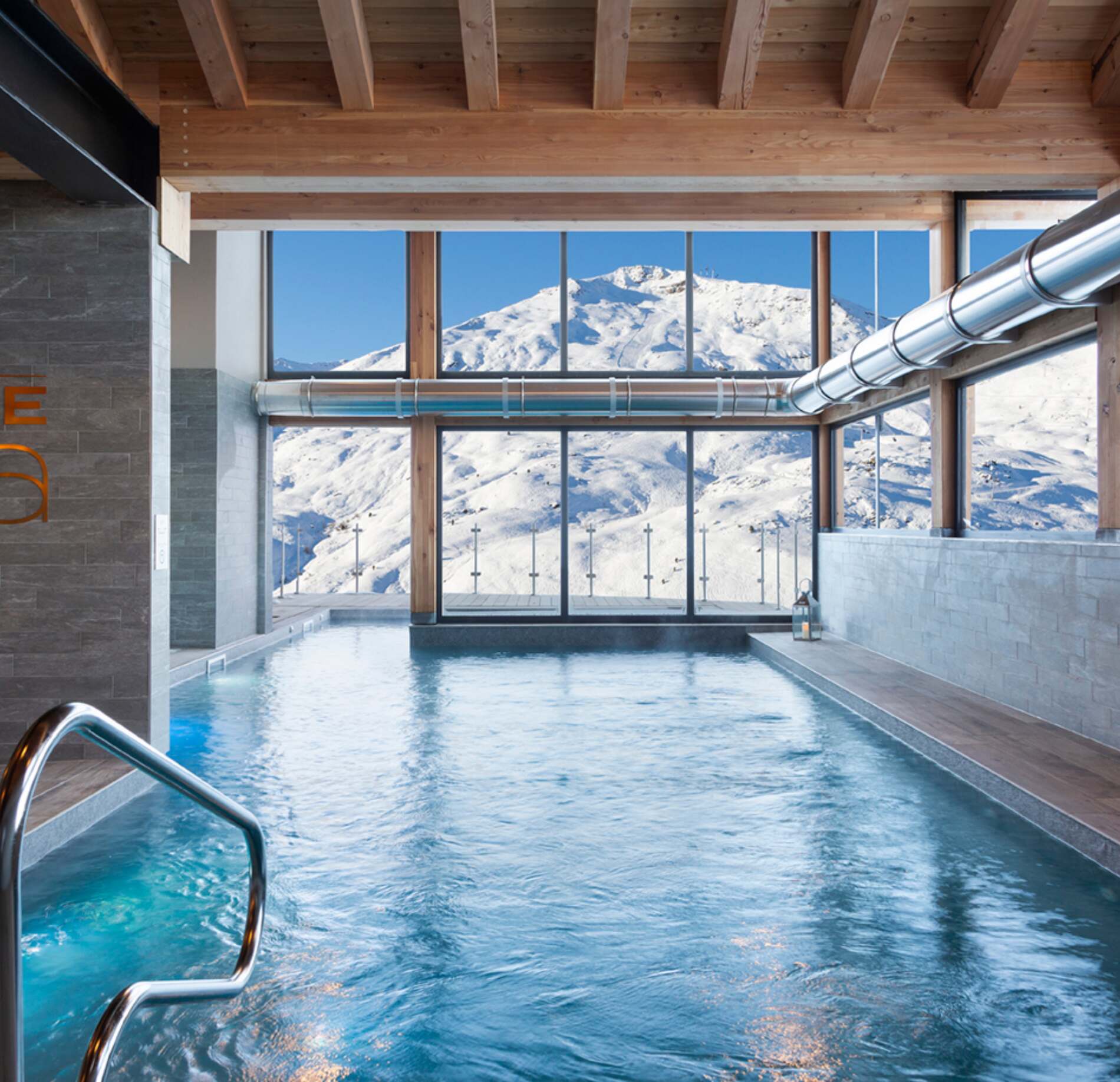 Une piscine intérieure du spa de l'hotel l'Ours Blanc s’étend sous une charpente en bois, face à de grandes baies vitrées donnant sur des montagnes enneigées.
L’eau légèrement agitée reflète la lumière naturelle, tandis qu’une échelle métallique et un bord en pierre complètent cet espace lumineux et apaisant.
