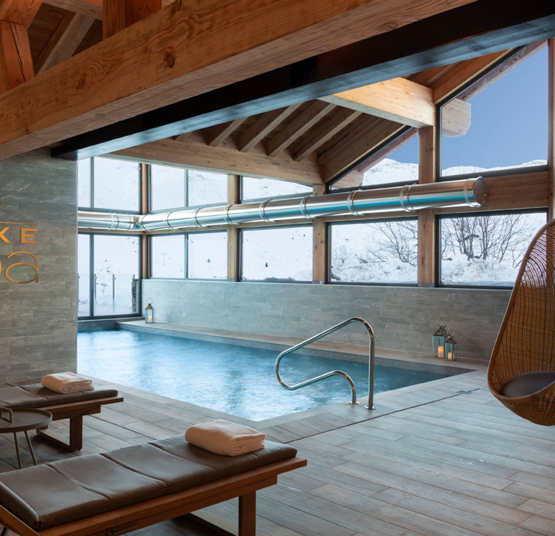Une piscine intérieure du spa de l'hotel l'Ours Blanc s’étend dans une pièce lumineuse avec une charpente en bois apparente et de grandes baies vitrées.
Des transats avec serviettes sont disposés au bord du bassin, tandis que la vue donne sur un paysage enneigé.