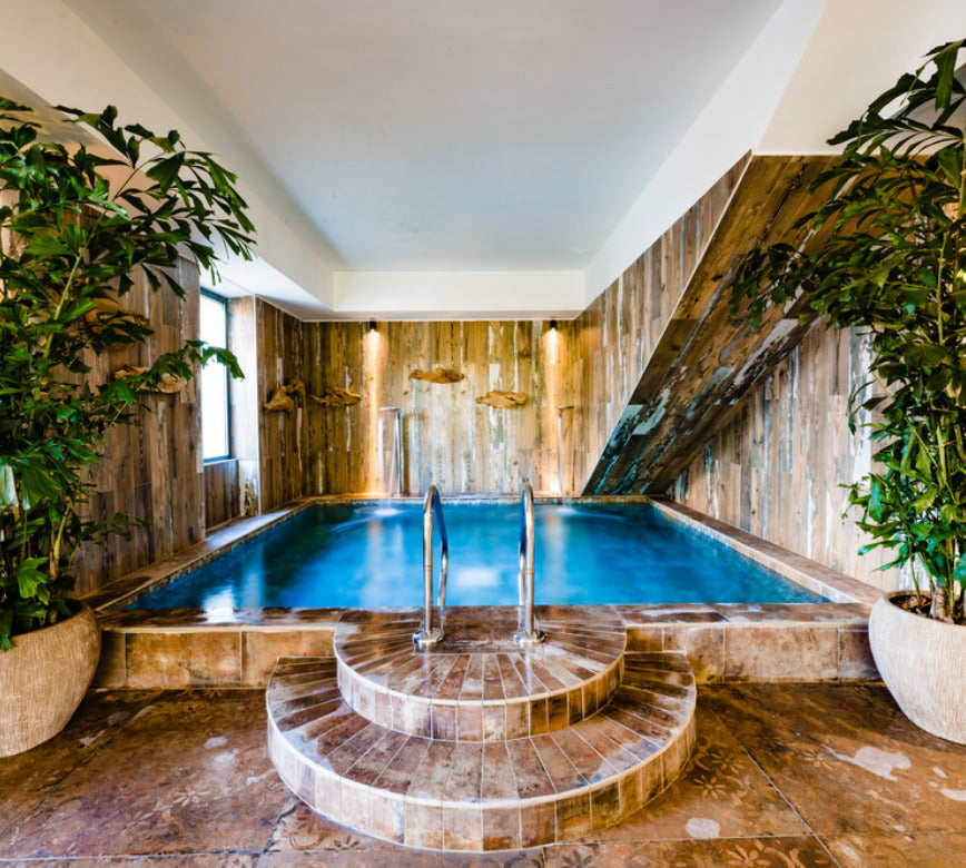 Espace aquatique du spa NUXE à l’Hôtel Les Pesquiers, composé d’un bassin intérieur encadré de bois brut, d’un accès par marches arrondies en pierre, de plantes décoratives et d’un éclairage doux valorisant les textures naturelles, offrant un cadre raffiné, enveloppant et propice à la détente
