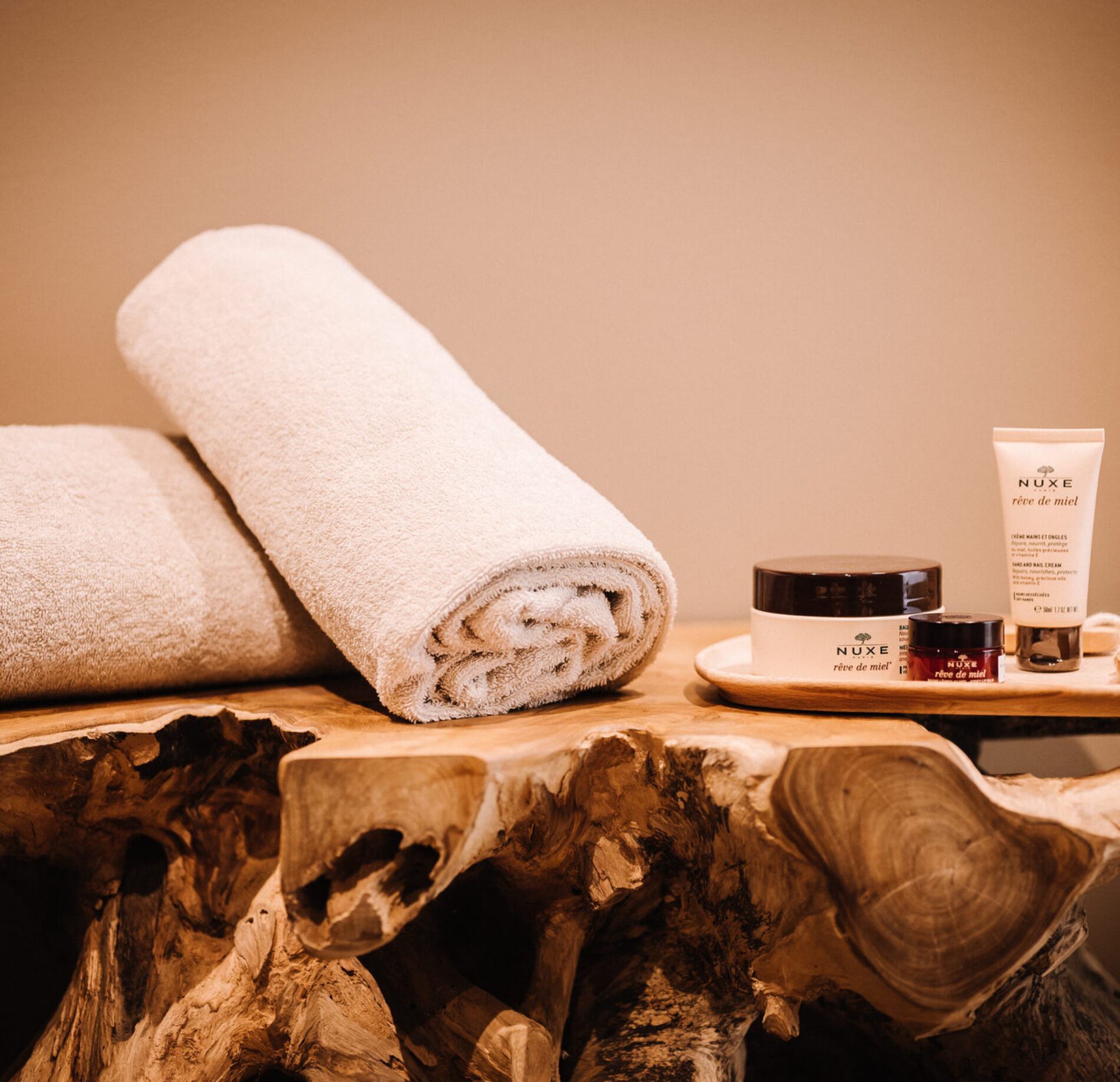 Détail de produits de soin du spa NUXE à l’Hôtel Les Pesquiers, avec serviettes douces soigneusement roulées et cosmétiques disposés sur un support en bois naturel sculpté, dans une atmosphère chaleureuse, élégante et apaisante.