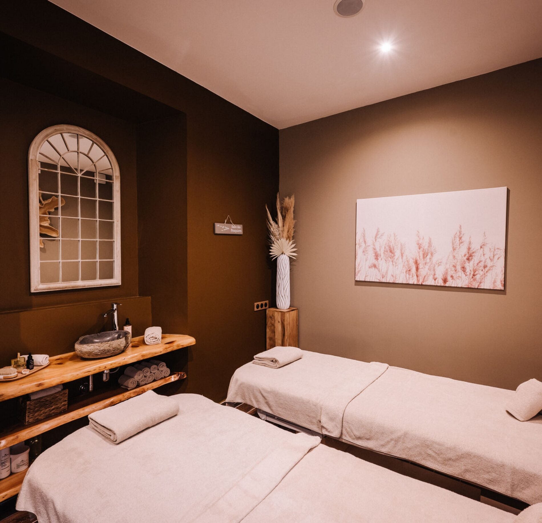 Cabine de soin chaleureuse du spa NUXE à l’Hôtel Les Pesquiers, avec deux tables de massage habillées de linge clair, éclairage doux, mobilier en bois naturel et décoration épurée créant une atmosphère intime, apaisante et raffinée. 