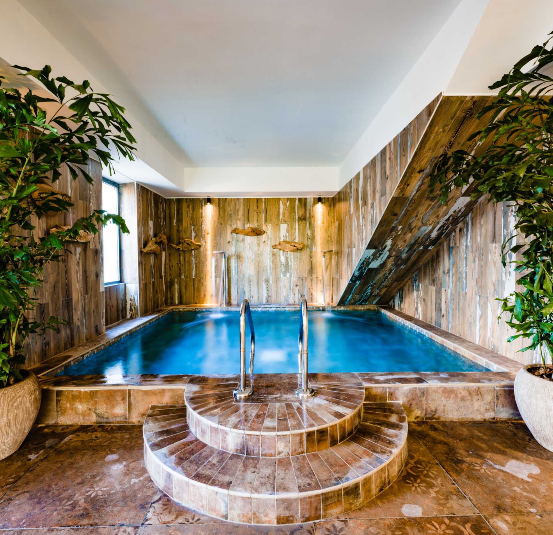 Piscine intérieure élégante du spa NUXE à l’Hôtel Les Pesquiers, avec bassin aux reflets turquoise entouré de murs en bois naturel, escalier circulaire en pierre et végétation luxuriante, créant une atmosphère chaleureuse, immersive et apaisante.