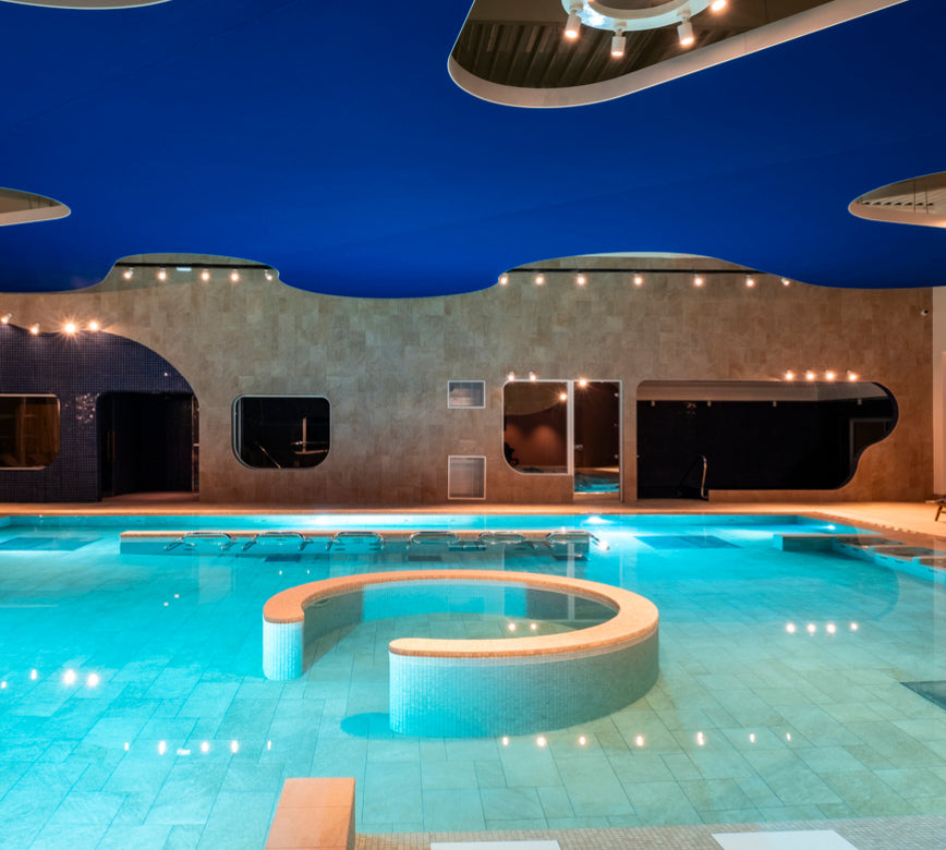 Une piscine intérieure élégante du Quiberon Mercure hotel & spa, avec une eau turquoise claire et des formes arrondies au centre du bassin pour la détente.
Le décor moderne se distingue par un plafond ondulé bleu intense et des ouvertures murales aux formes organiques, éclairées par une lumière chaude et apaisante.