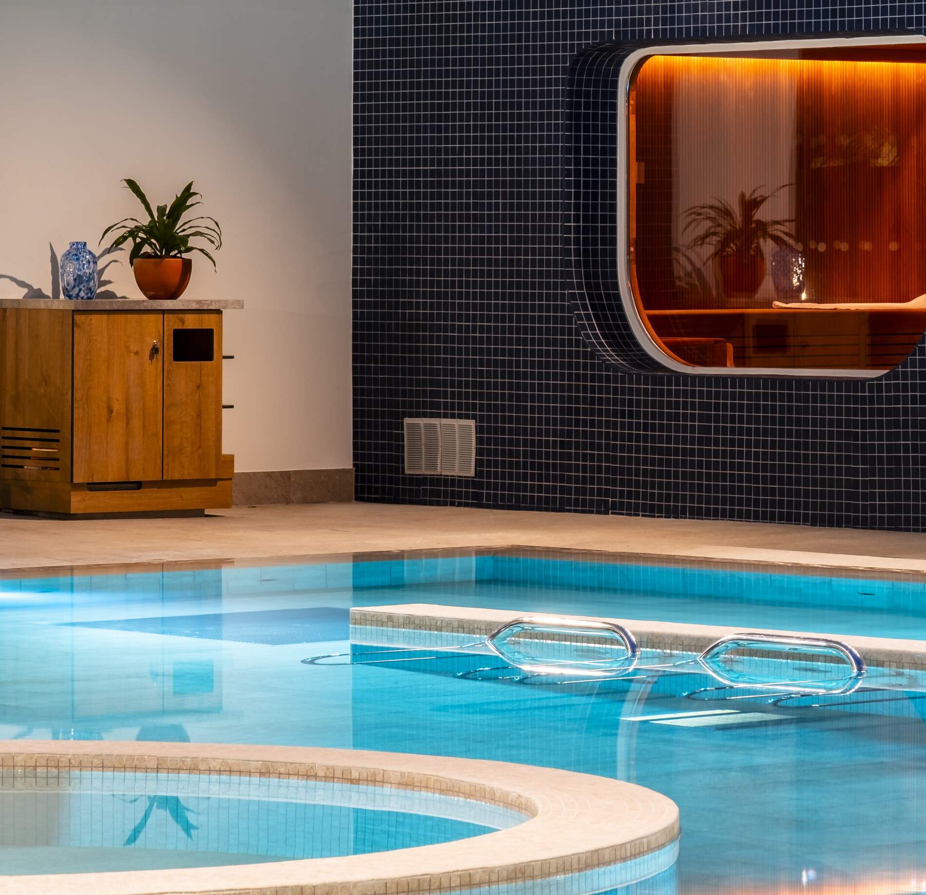 il y a une piscine intérieure élégante du Quiberon Mercure hotel & spa, avec une eau turquoise limpide et des marches immergées accompagnées de rampes métalliques.
À côté du bassin, un mur en mosaïque sombre encadre une ouverture lumineuse orangée, tandis qu’un petit meuble en bois et une plante apportent une touche chaleureuse.