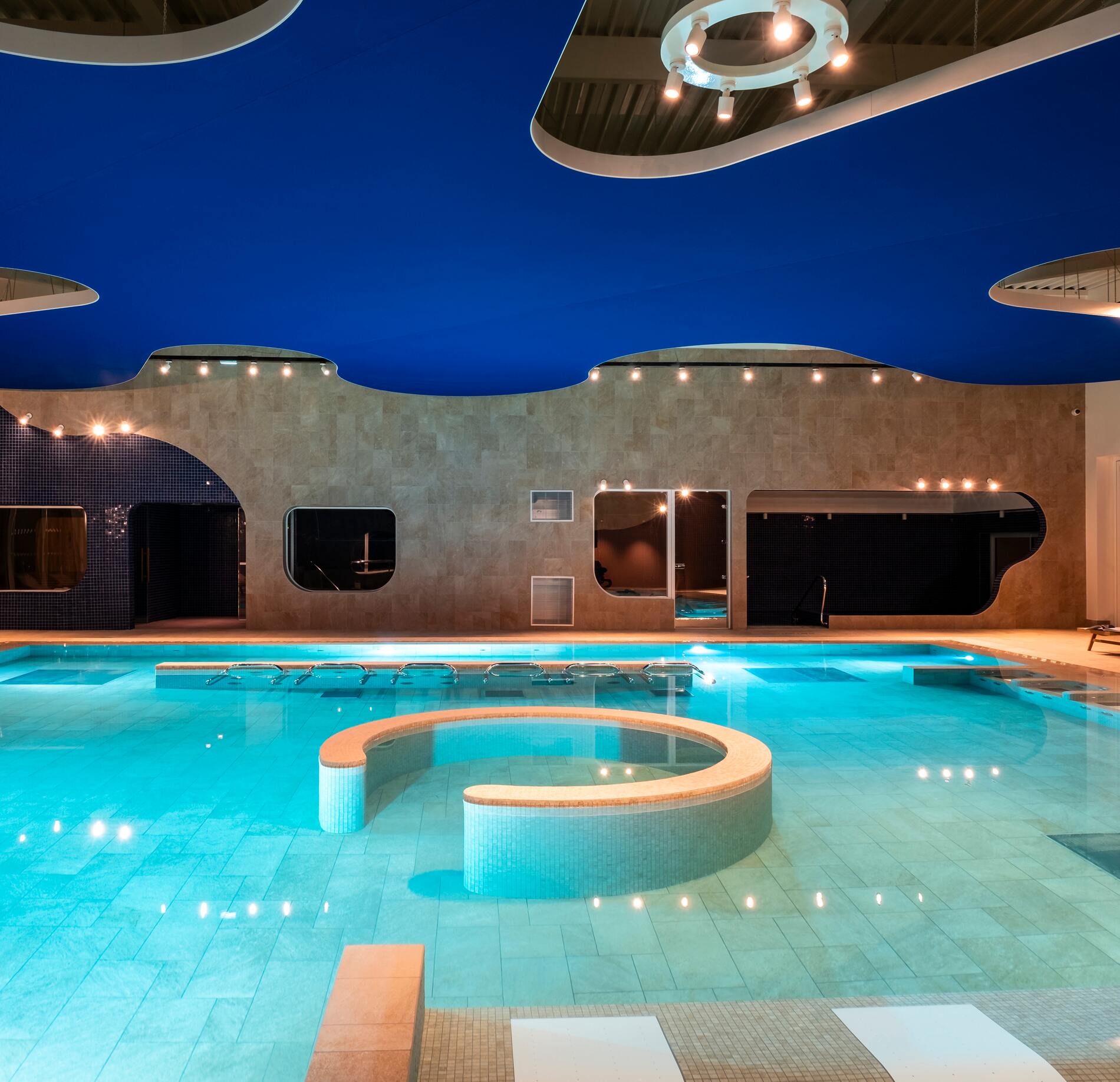 Il y a une piscine intérieure moderne du Quiberon Mercure hotel & spa, avec une eau turquoise limpide et des formes courbes dédiées à la relaxation.
L’ambiance contemporaine se distingue par un plafond ondulé bleu profond et des ouvertures murales organiques baignées d’une lumière douce et chaleureuse.