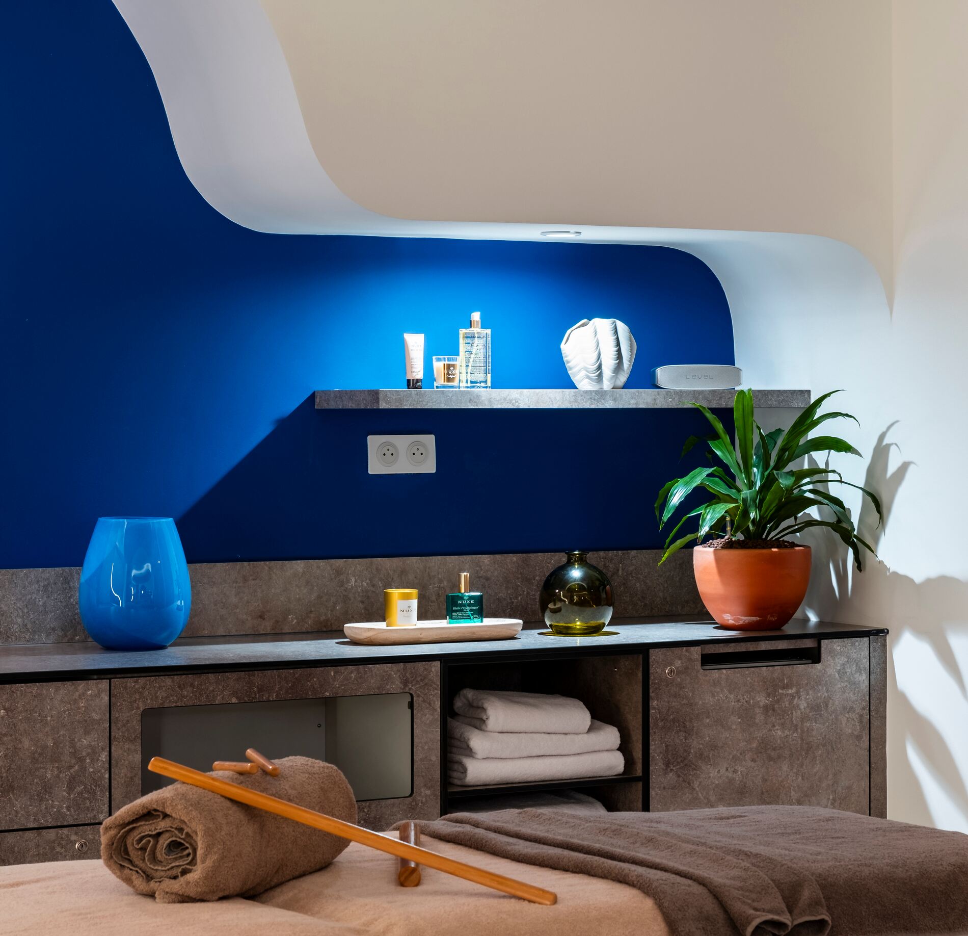 Il y a un espace de soin apaisant du Quiberon Mercure hotel & spa, avec une table de massage recouverte de serviettes et des accessoires de bien-être soigneusement disposés.
Le décor mêle un mur bleu profond intégré dans des formes arrondies, des étagères avec produits et une plante verte apportant une ambiance relaxante et chaleureuse.