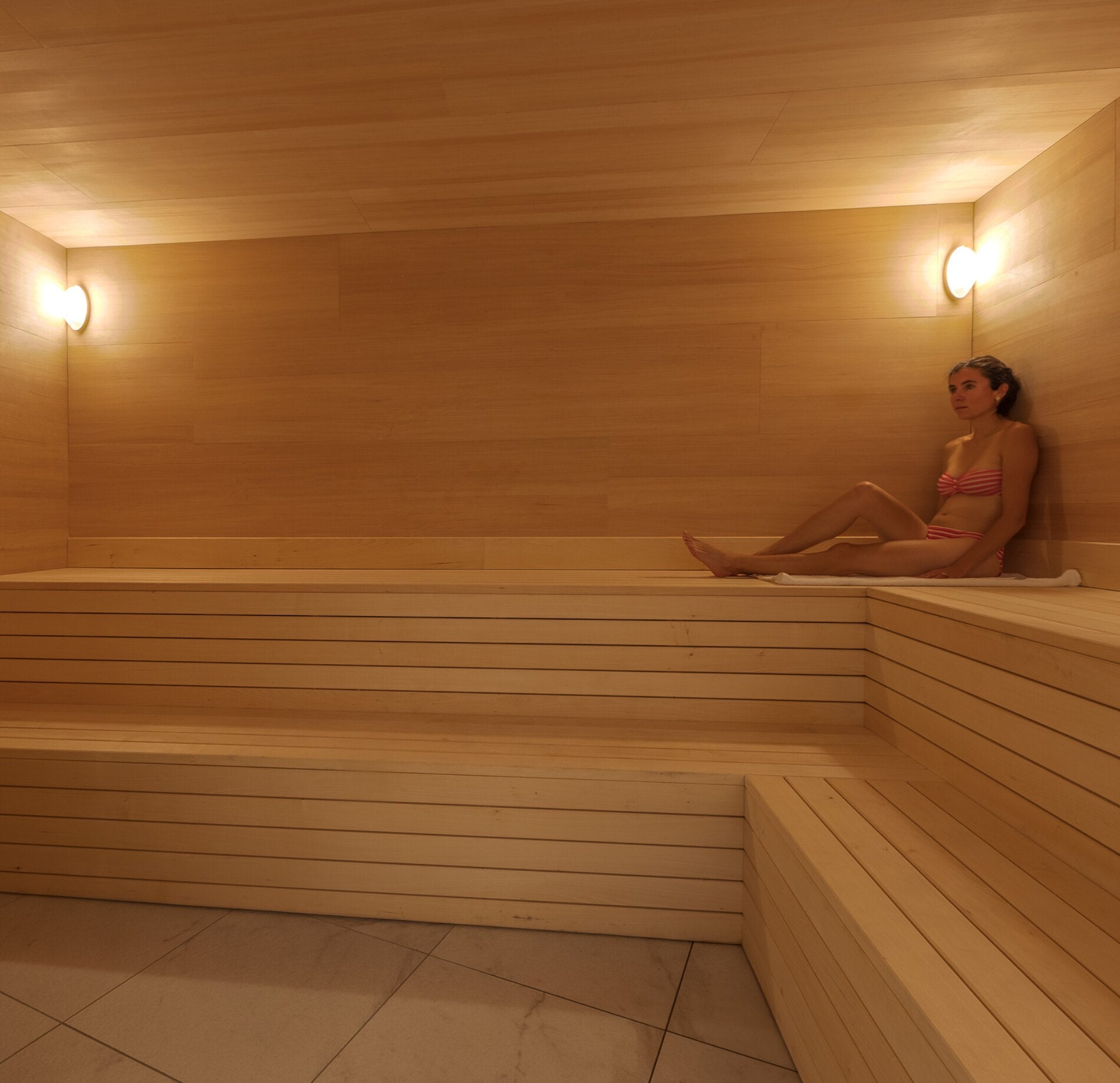 Un sauna en bois chaleureux du Rivage Hotel Spa, avec des bancs en lattes claires disposés en gradins sur plusieurs niveaux.
Une personne assise sur le côté profite de la chaleur dans une ambiance douce, éclairée par des appliques murales diffusant une lumière chaleureuse et enveloppante.