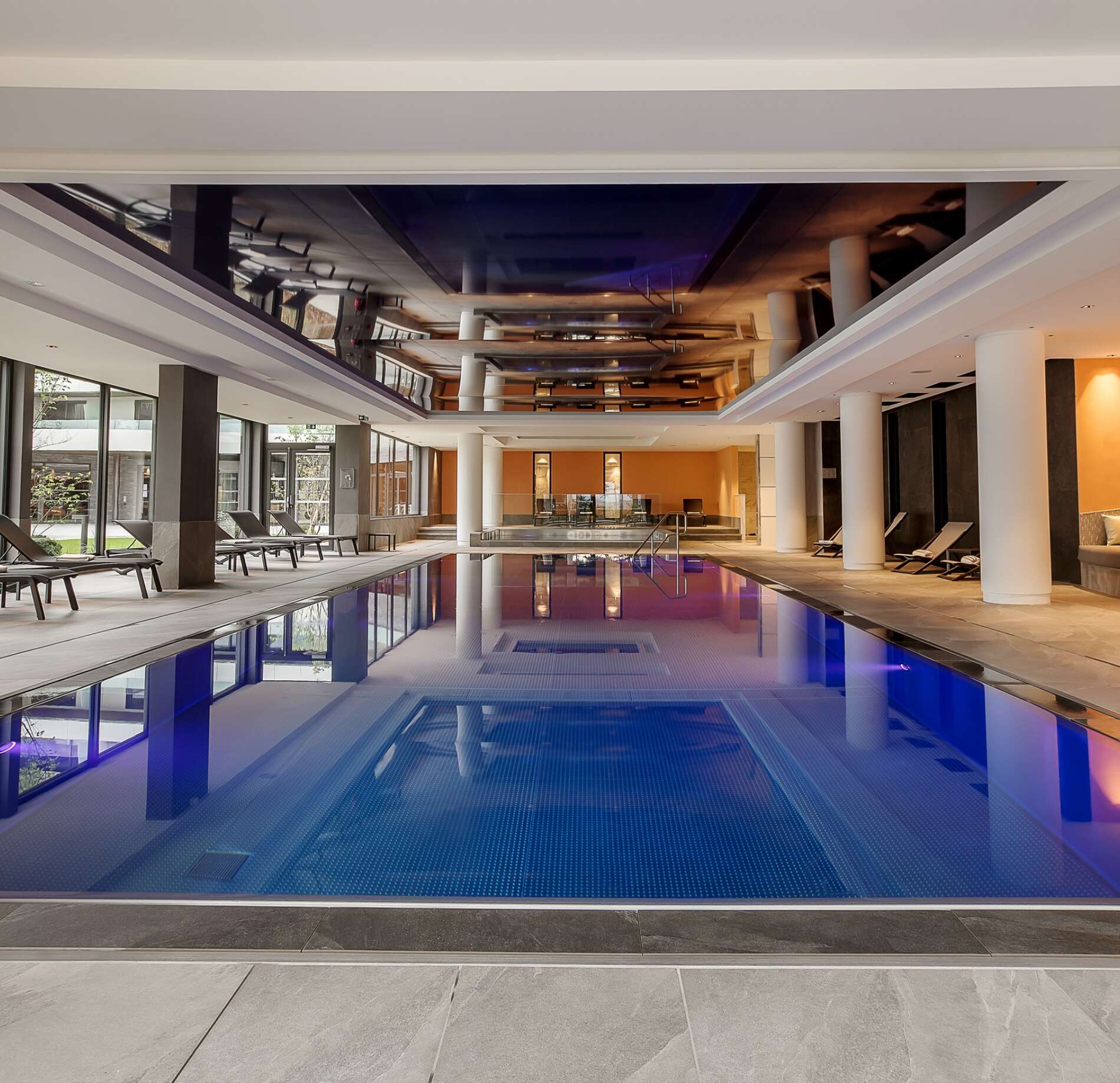 Une piscine intérieure spacieuse du Rivage Hotel Spa, avec un grand bassin aux reflets bleu profond entouré de colonnes blanches et de larges baies vitrées.
Le plafond miroir et l’éclairage varié créent une ambiance moderne et immersive, tandis que des transats alignés sur les côtés invitent à la détente.