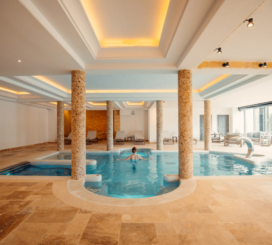 Grand bassin de bien-être au spa NUXE, structuré autour de colonnes minérales et éclairé par un plafond aux lignes lumineuses, avec zones de relaxation aménagées de transats confortables et larges baies vitrées, offrant une ambiance calme et haut de gamme.