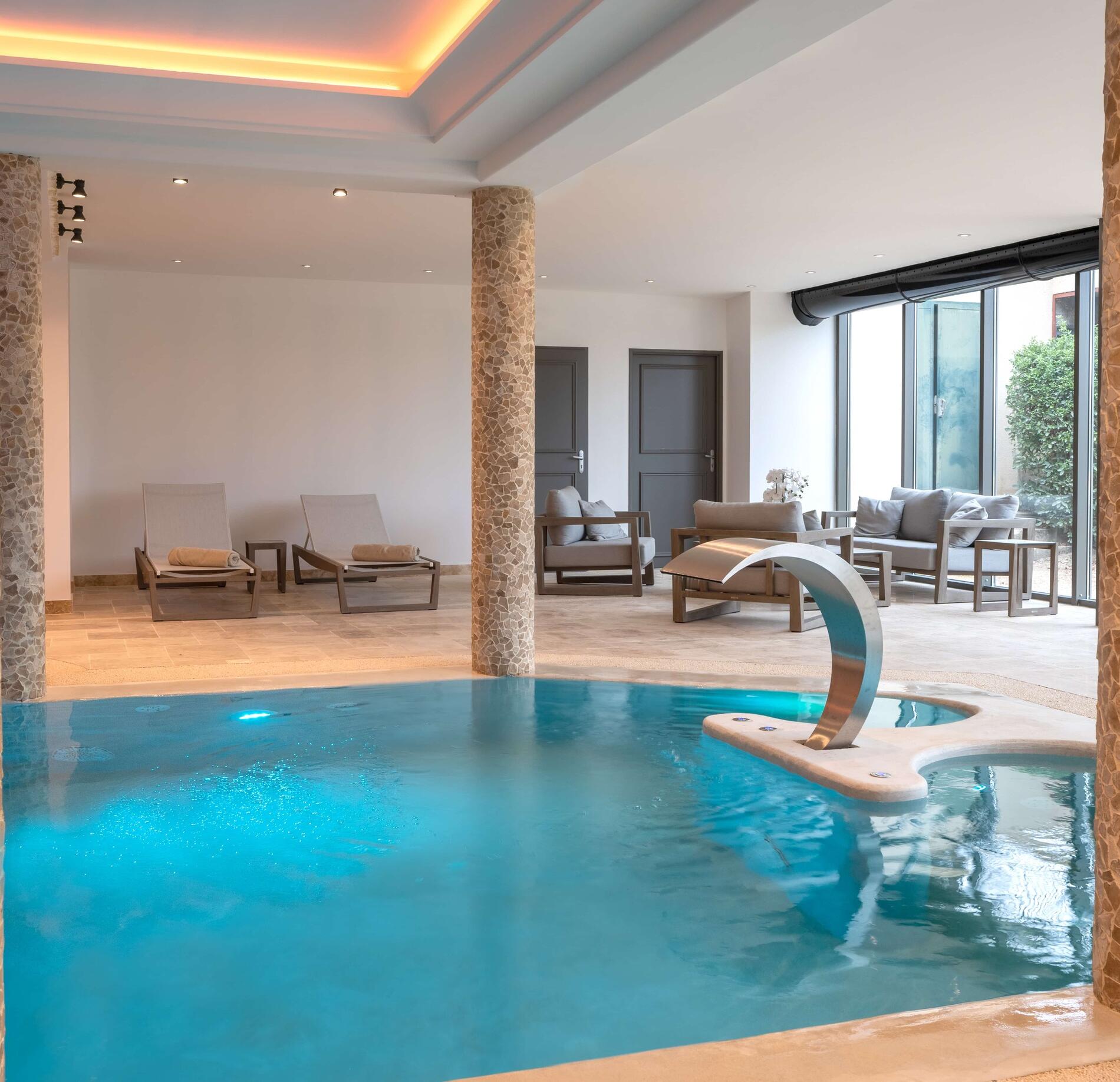 Piscine intérieure élégante du spa NUXE, avec eau turquoise lumineuse et fontaine moderne en inox, entourée de transats confortables et baies vitrées offrant une vue apaisante sur l’extérieur, dans une atmosphère raffinée et relaxante. 