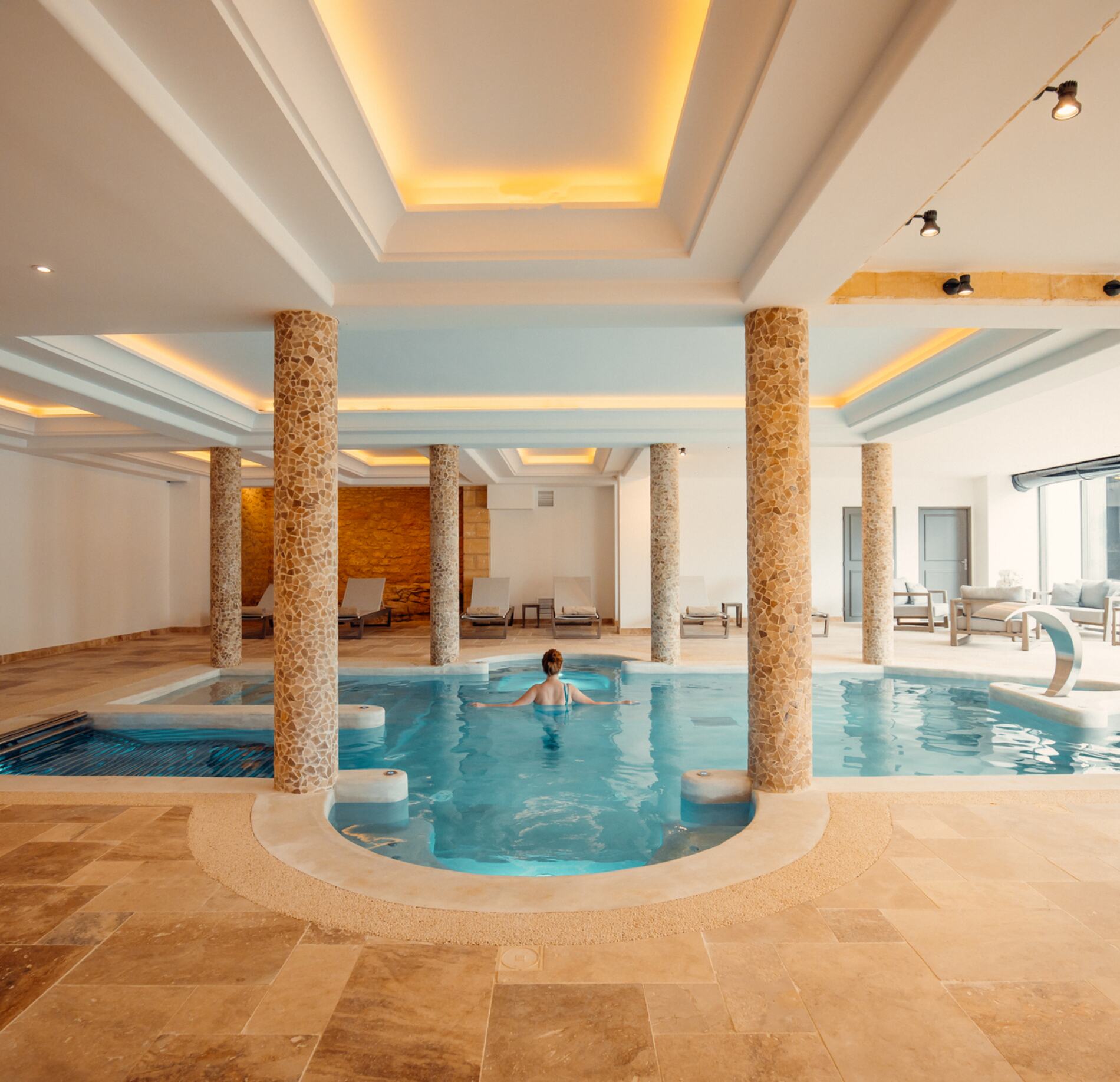 Vaste piscine intérieure du spa NUXE, entourée de colonnes en pierre et baignée d’une lumière douce, avec eau turquoise apaisante, espace détente équipé de transats élégants et architecture lumineuse créant une atmosphère raffinée et sereine. 
