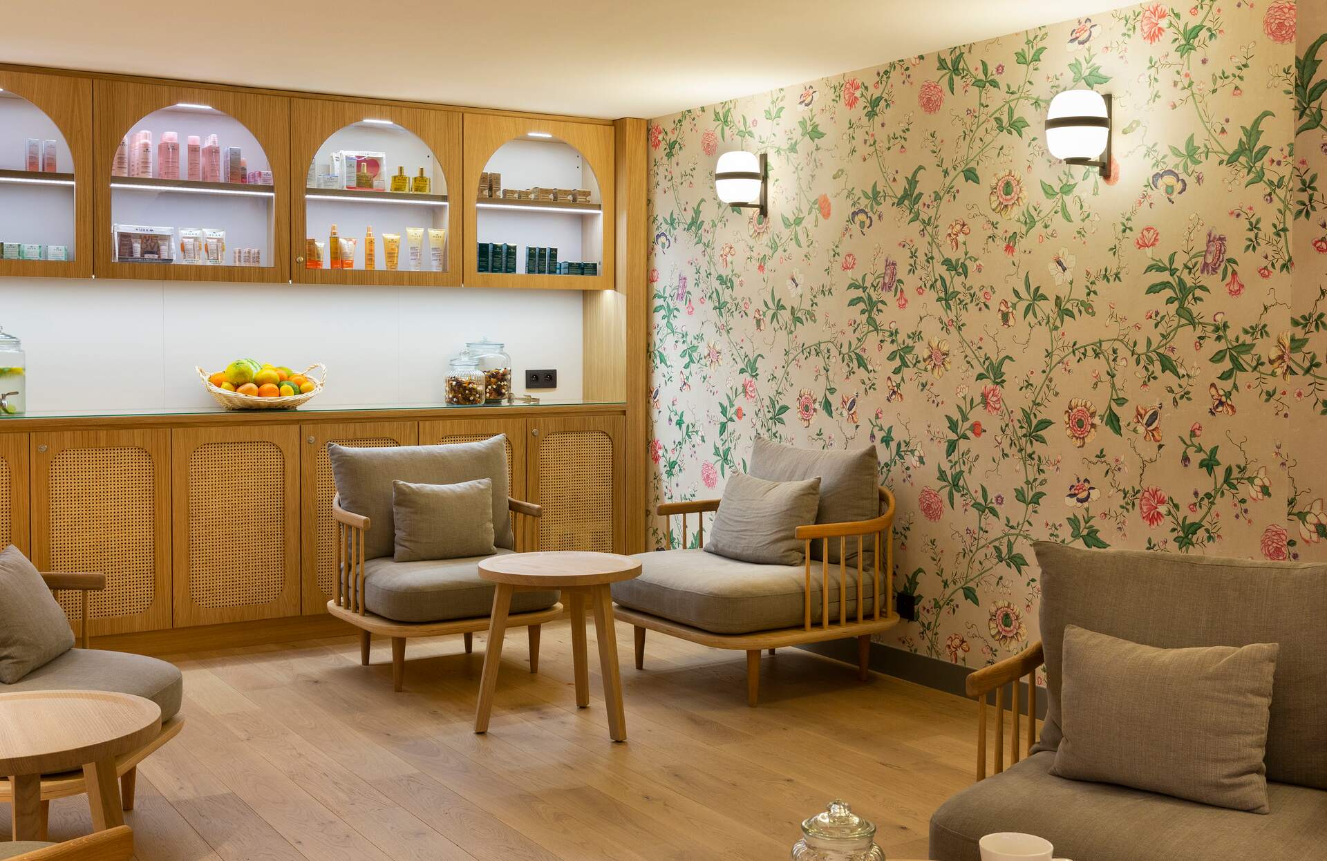 Un espace de détente chaleureux s’organise autour de fauteuils en bois aux lignes arrondies et de coussins confortables, disposés autour de petites tables basses.
À l’arrière, des étagères intégrées présentent des produits et des éléments décoratifs, avec un mur à motif floral et un éclairage doux qui créent une ambiance élégante et apaisante.