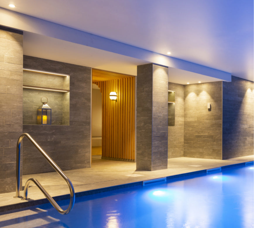 Une piscine intérieure élégante de l’hotel spa Rocky Pop, avec une eau bleutée et un escalier métallique plongeant dans le bassin.
L’espace est entouré de murs en pierre grise et d’une ouverture en bois lumineux menant à une zone de détente, éclairée par des lumières douces et chaleureuses.