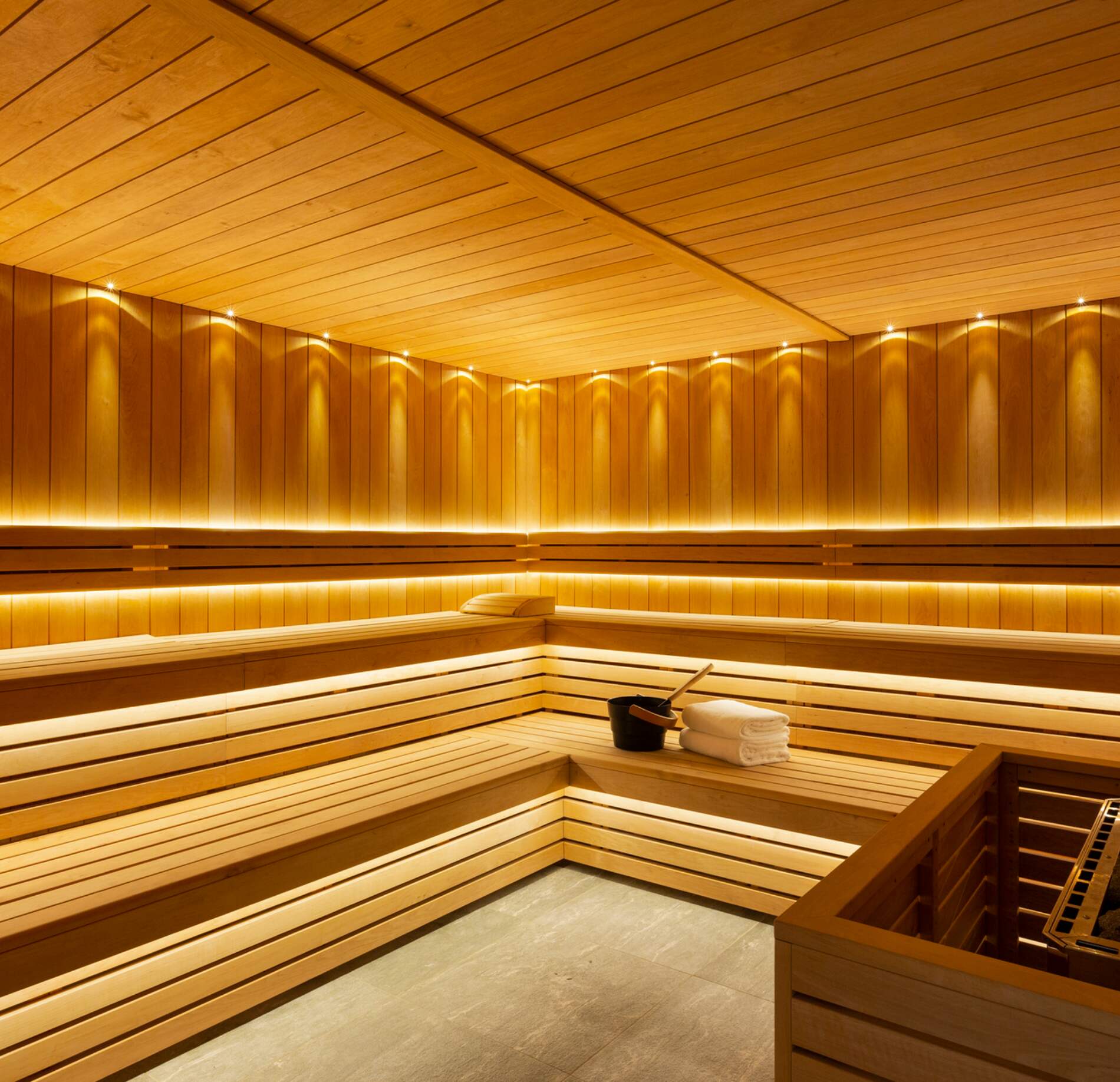 Un sauna en bois chaleureux de l’hotel spa Rocky Pop, entièrement habillé de lattes dorées avec des bancs disposés en gradins sur plusieurs niveaux.
Un éclairage indirect souligne les lignes du bois et crée une ambiance enveloppante, avec des serviettes pliées et un seau posé au centre pour les rituels traditionnels.