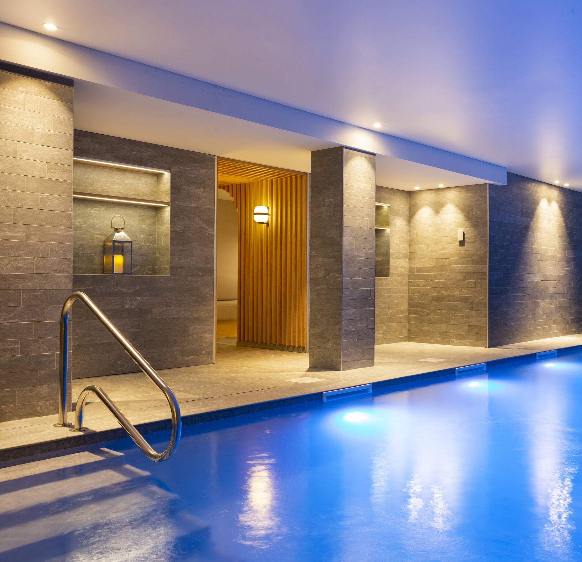 Une piscine intérieure élégante de l’hotel spa Rocky Pop, avec une eau bleu clair et un escalier métallique menant dans le bassin.
L’espace est entouré de murs en pierre grise et d’une ouverture en bois clair donnant sur une zone de repos, éclairée par des lumières douces et cosy.
