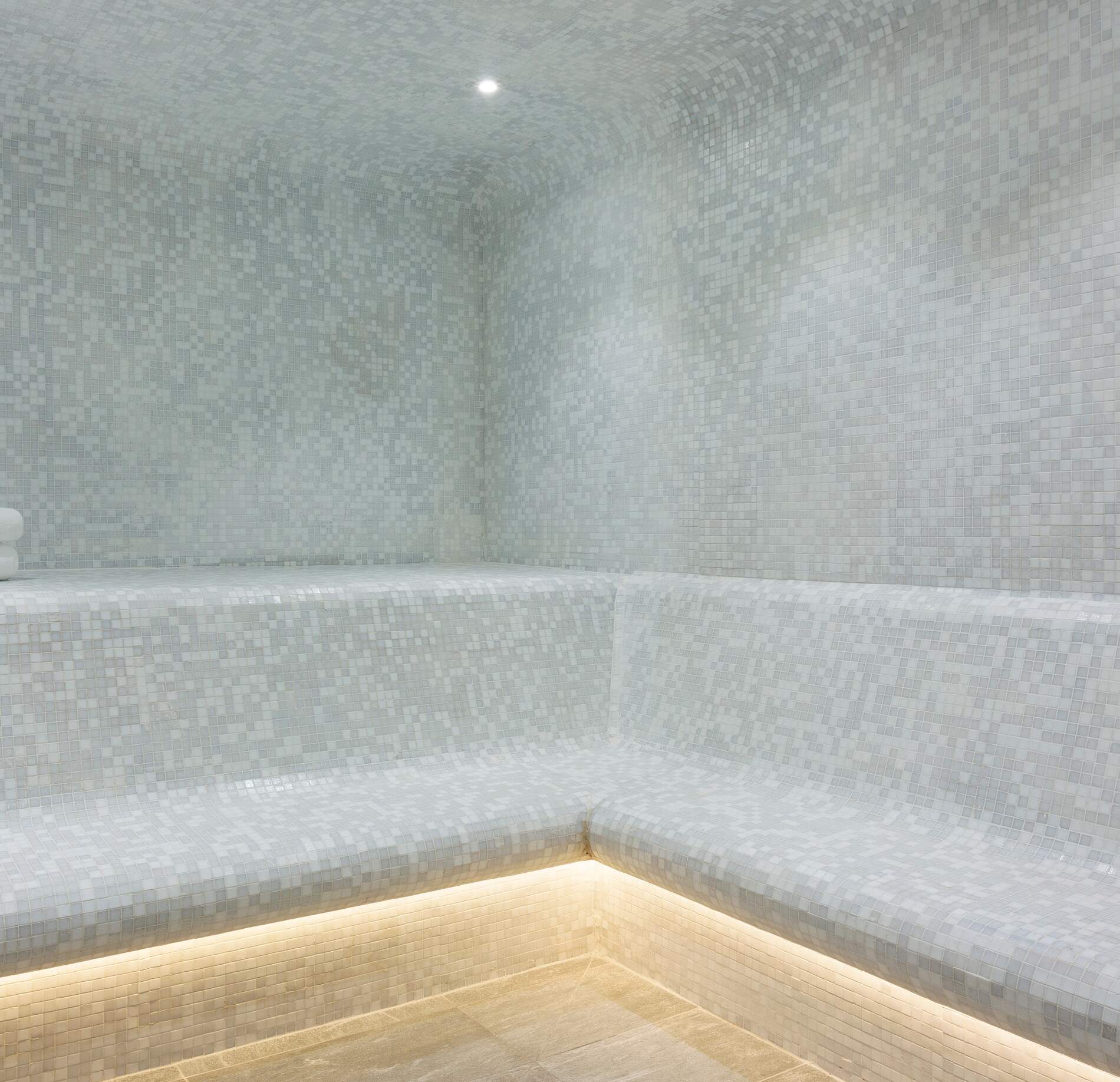 Un hammam minéral et apaisant de l’hotel spa Rocky Pop, entièrement recouvert de petites mosaïques claires formant des assises en angle.
La lumière douce diffusée depuis le plafond et sous les bancs enveloppe l’espace d’une ambiance feutrée, propice à la détente et au calme.