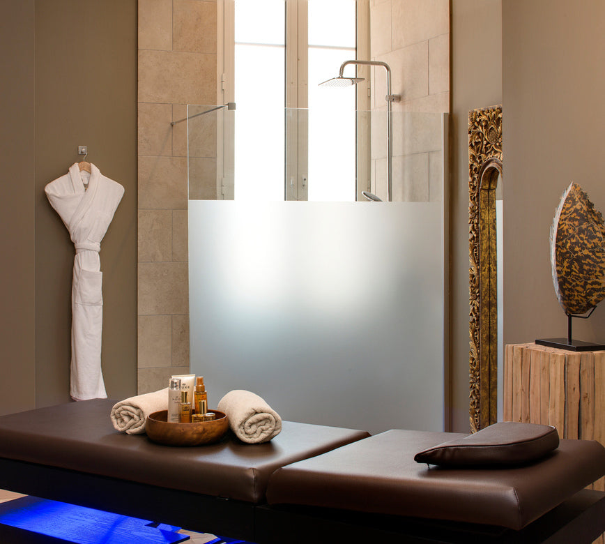 Une cabine de soins du spa du Grand Hotel des Sablettes présente une table de massage en cuir foncé avec des serviettes roulées et des produits disposés au centre.
En arrière-plan, une douche vitrée devant une grande fenêtre diffuse une lumière douce, tandis qu’un peignoir suspendu et des éléments décoratifs créent une ambiance élégante et apaisante.
