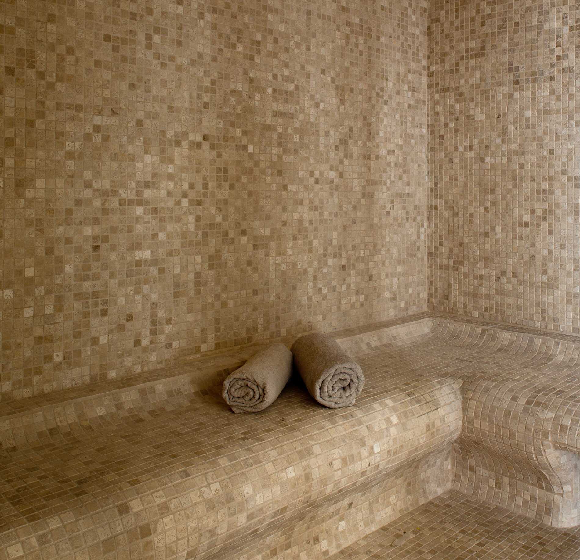 Un hammam du spa du Grand Hotel des Sablettes est entièrement recouvert de petites mosaïques beiges, avec des bancs intégrés aux formes arrondies.
Deux serviettes roulées sont posées sur le banc, tandis que l’ensemble de la pièce crée une ambiance douce, uniforme et apaisante.