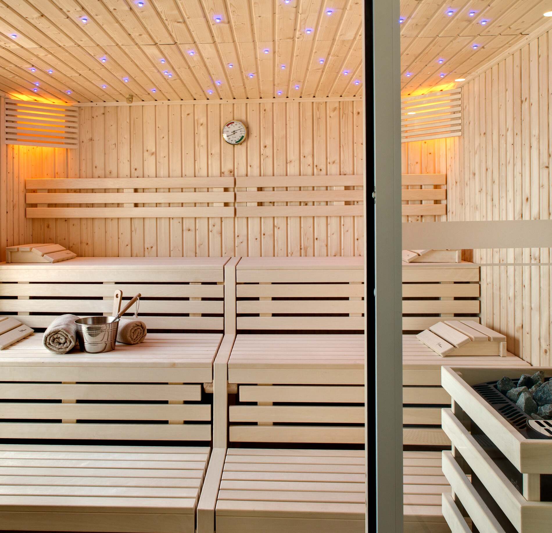 Un sauna du spa du Grand Hotel des Sablettes entièrement en bois clair présente plusieurs bancs superposés avec des serviettes et un seau avec louche posés dessus.
Le plafond est parsemé de petites lumières bleutées, tandis qu’un éclairage chaud sur les côtés crée une ambiance à la fois apaisante et moderne