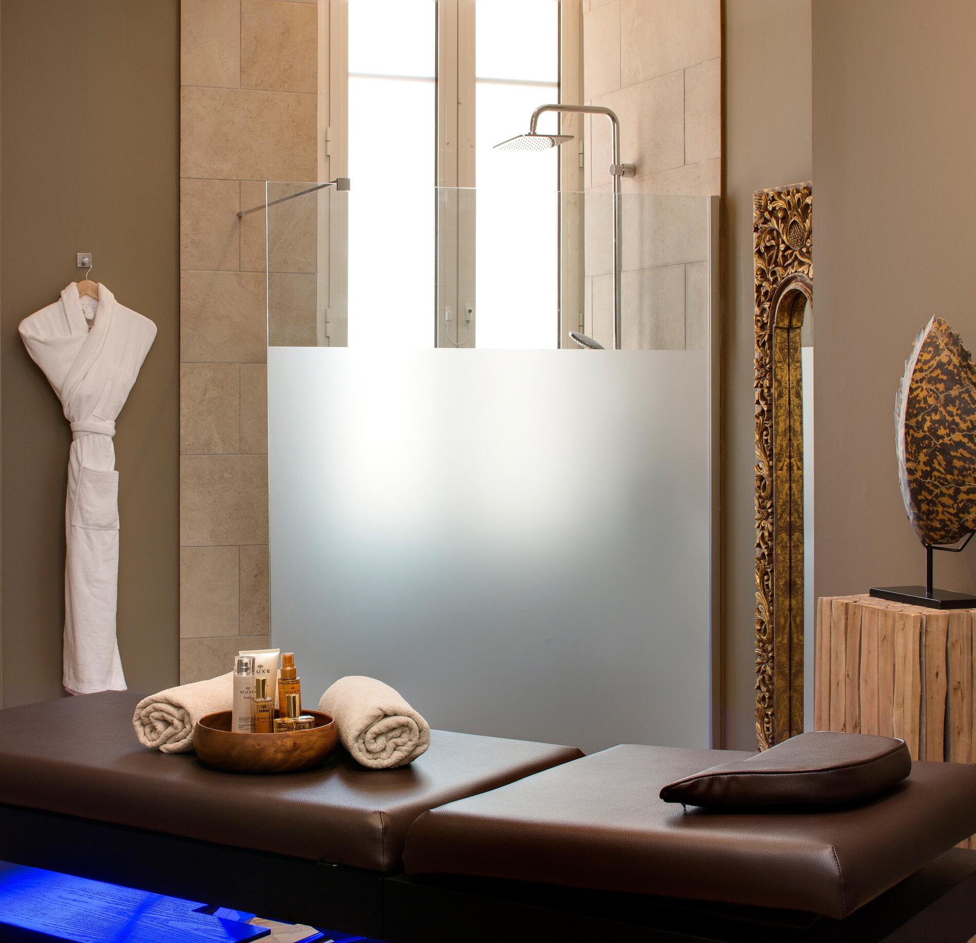 Une cabine de soins du spa du Grand Hotel des Sablettes présente une table de massage en cuir foncé avec des serviettes roulées et des produits NUXE disposés au centre.
En arrière-plan, une douche vitrée devant une grande fenêtre diffuse une lumière douce, tandis qu’un peignoir suspendu et des éléments décoratifs créent une ambiance élégante et apaisante.