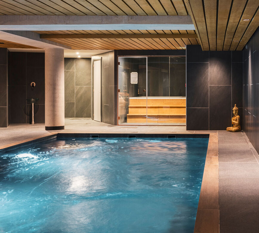 Espace bien-être du spa NUXE à Serre Chevalier, composé d’un bassin intérieur à l’eau animée, entouré de murs aux teintes minérales profondes, d’un plafond en bois aux lignes graphiques et d’un sauna vitré éclairé chaleureusement, avec statue décorative et éclairage tamisé offrant un cadre élégant, apaisant et propice à la détente.