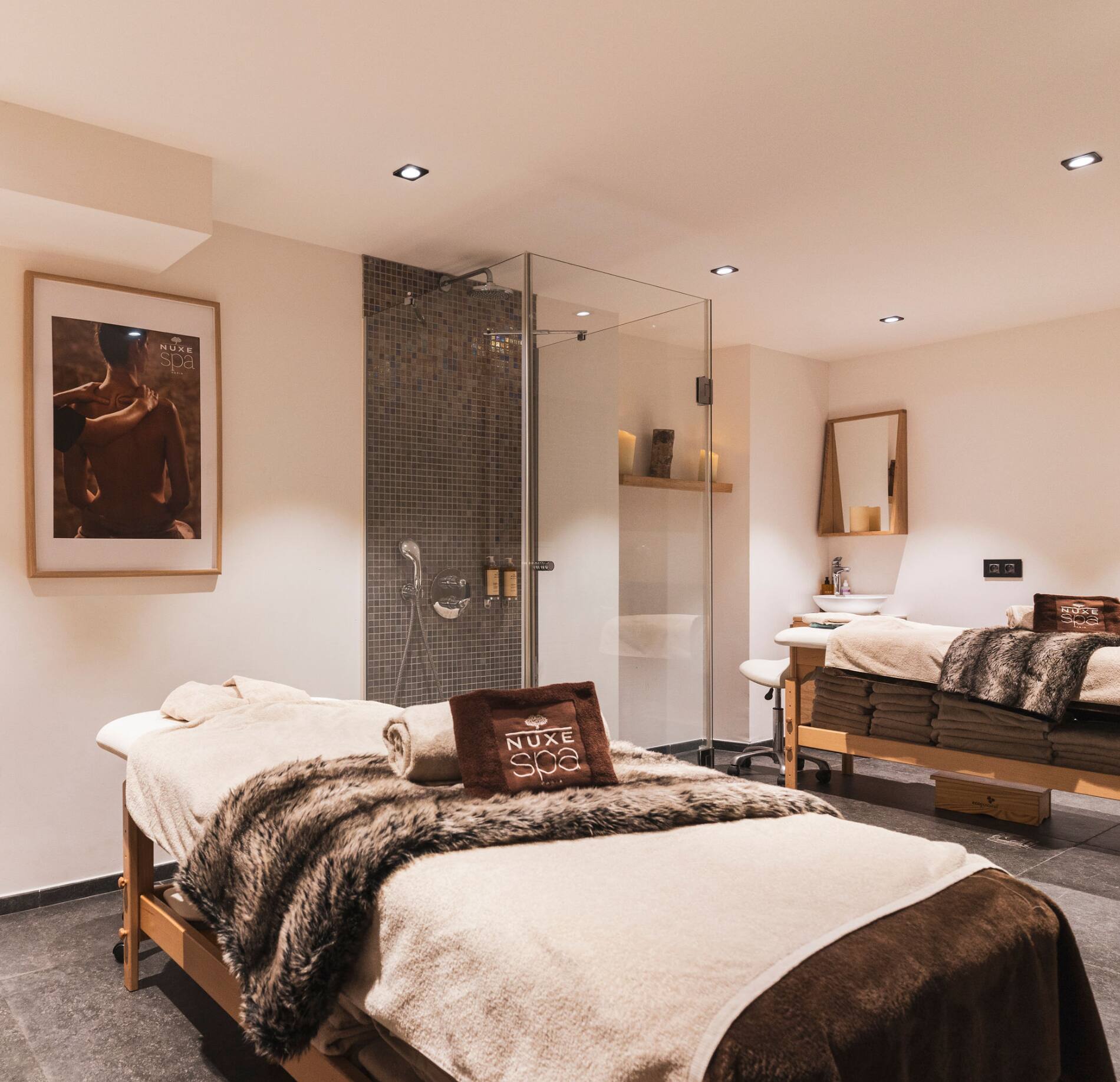 Cabine de soin élégante du spa NUXE à Serre Chevalier, avec deux tables de massage habillées de linge doux et plaids chaleureux, éclairage tamisé, douche contemporaine vitrée et décor épuré, créant une atmosphère intime, raffinée et apaisante.