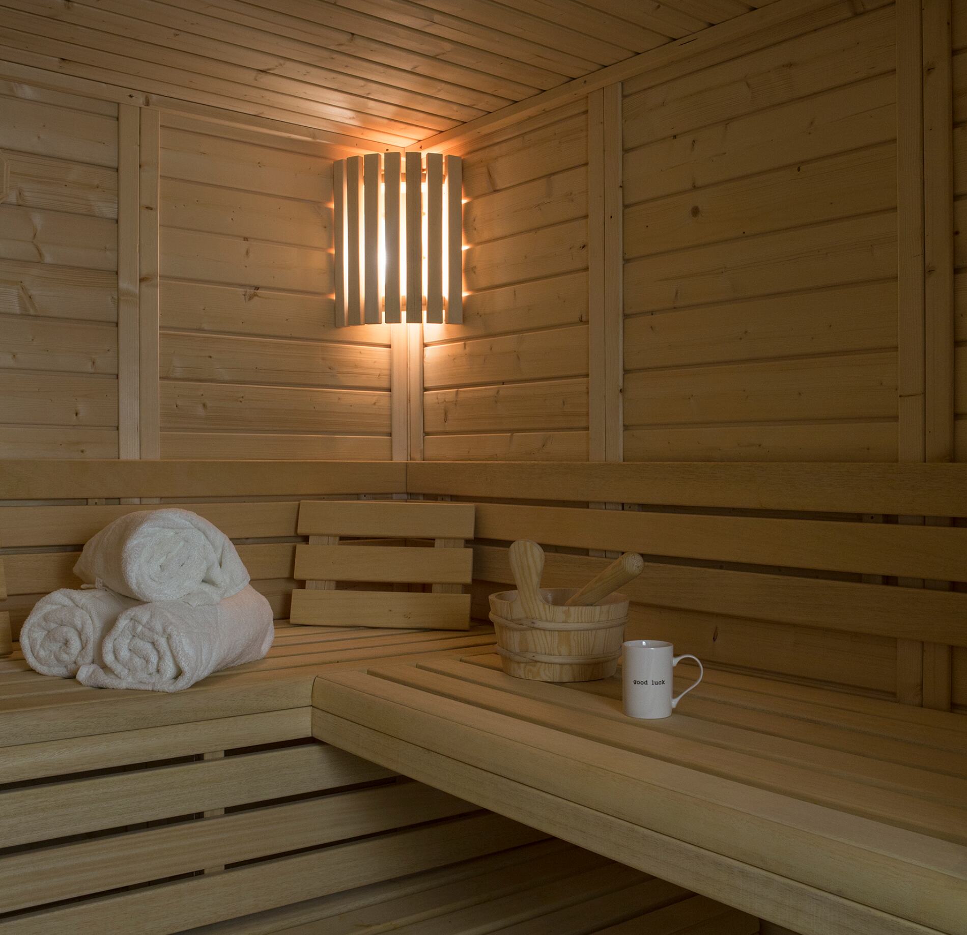 Sauna élégant du spa NUXE à Serre Chevalier, entièrement habillé de bois clair, avec banquettes spacieuses, serviettes roulées, seau traditionnel et éclairage mural chaleureux diffusant une lumière douce, créant une atmosphère intime et profondément relaxante. 