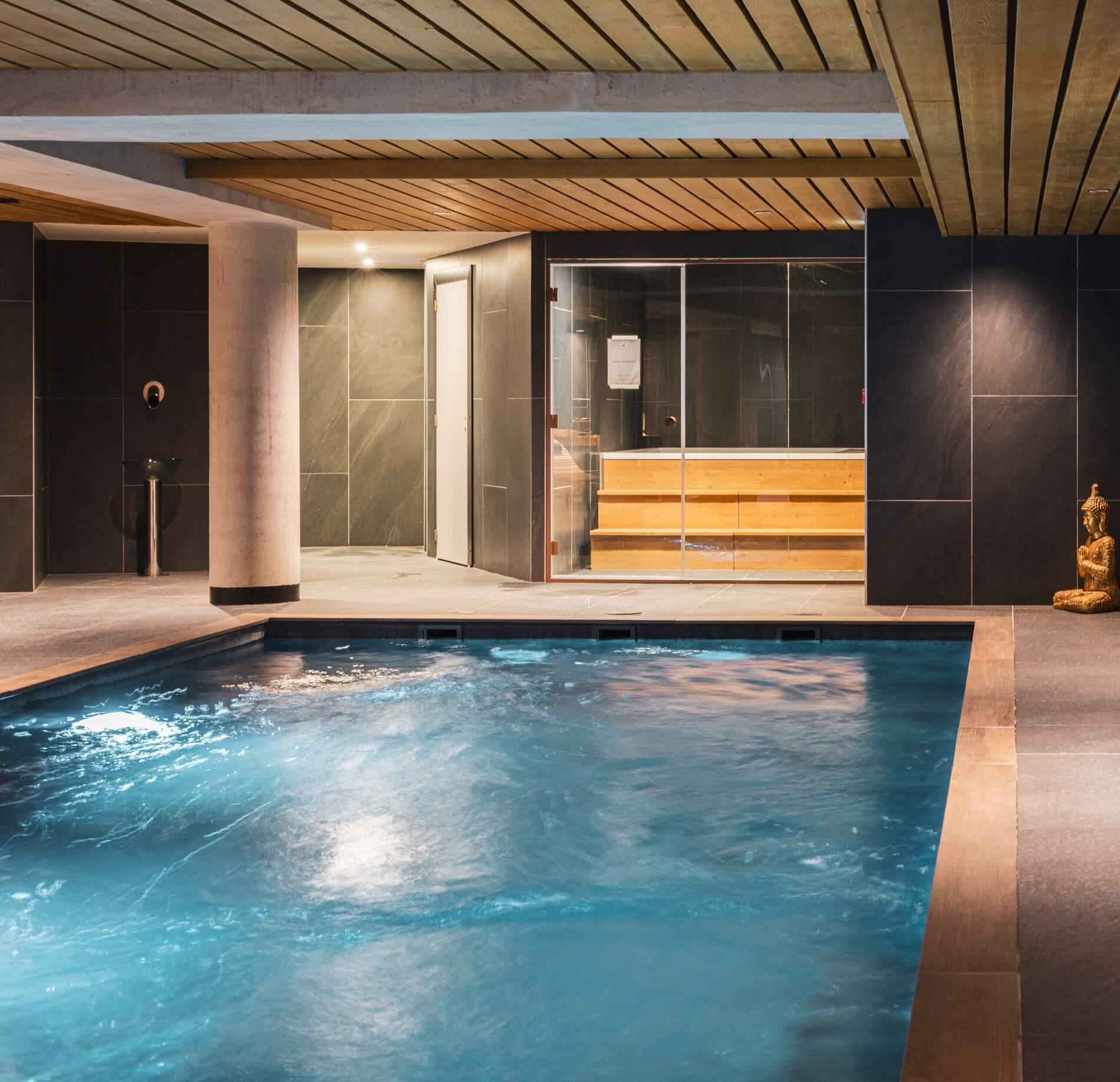 Piscine intérieure élégante du spa NUXE à Serre Chevalier, avec bassin aux remous apaisants, revêtements sombres raffinés, plafond en bois chaleureux et sauna vitré en arrière-plan, créant une atmosphère intime, contemporaine et haut de gamme.