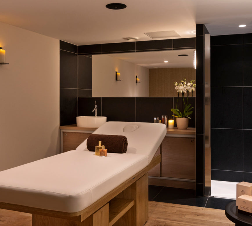 Une cabine de soin élégante du St'Alban Hotel & Spa, avec une table de massage recouverte d’un drap blanc, accompagnée de serviettes roulées et de flacons disposés avec soin.
Le décor associe des tons bois et noir, un grand miroir mural et un éclairage tamisé qui crée une atmosphère intime et apaisante.