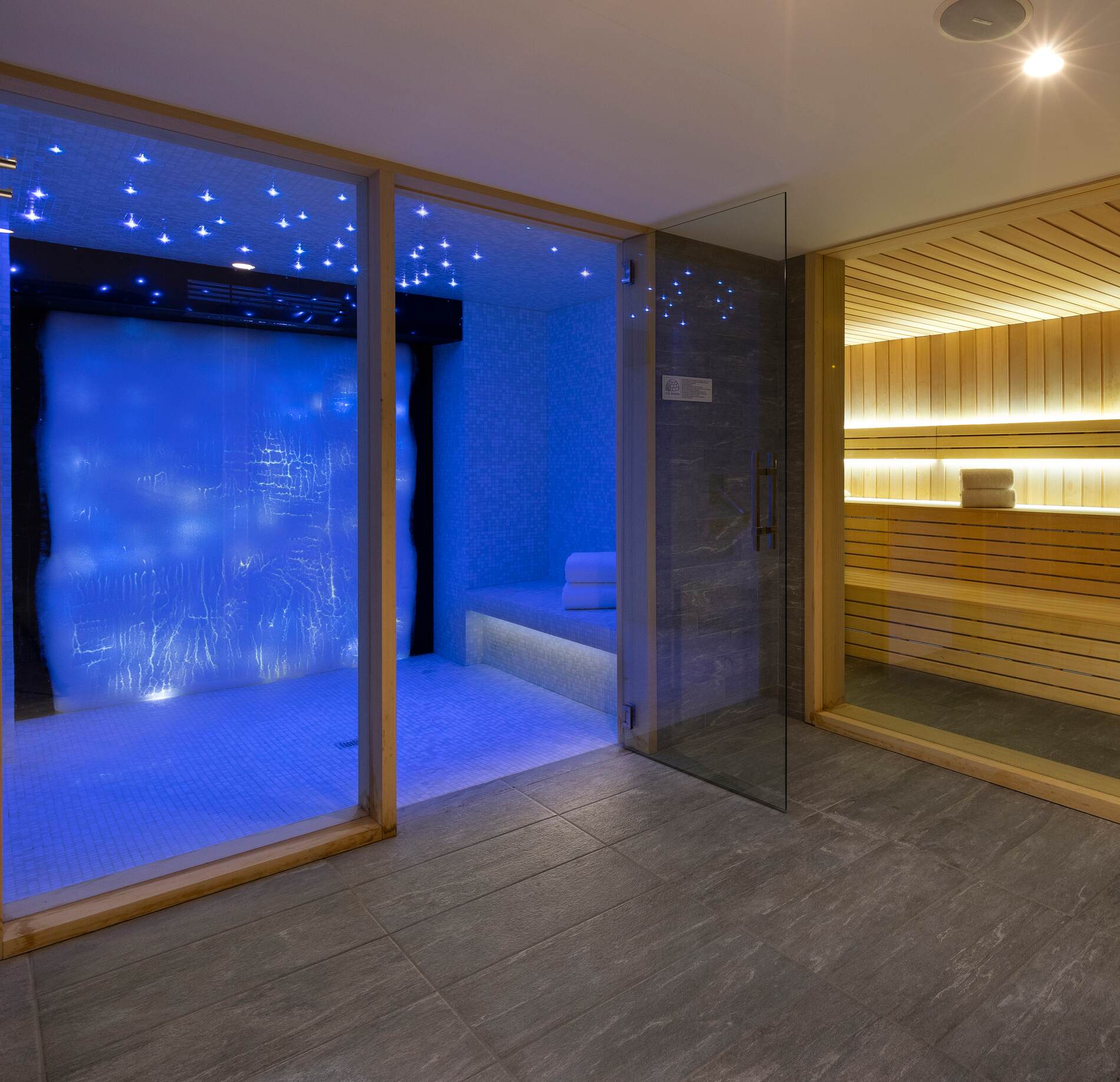 Un espace bien‑être moderne du St'Alban Hotel & Spa, où un hammam aux parois bleutées et au plafond étoilé diffuse une lumière douce et immersive.
À côté, un sauna en bois aux lignes épurées contraste avec la fraîcheur du hammam, séparé par une paroi vitrée dans une ambiance élégante et apaisante.