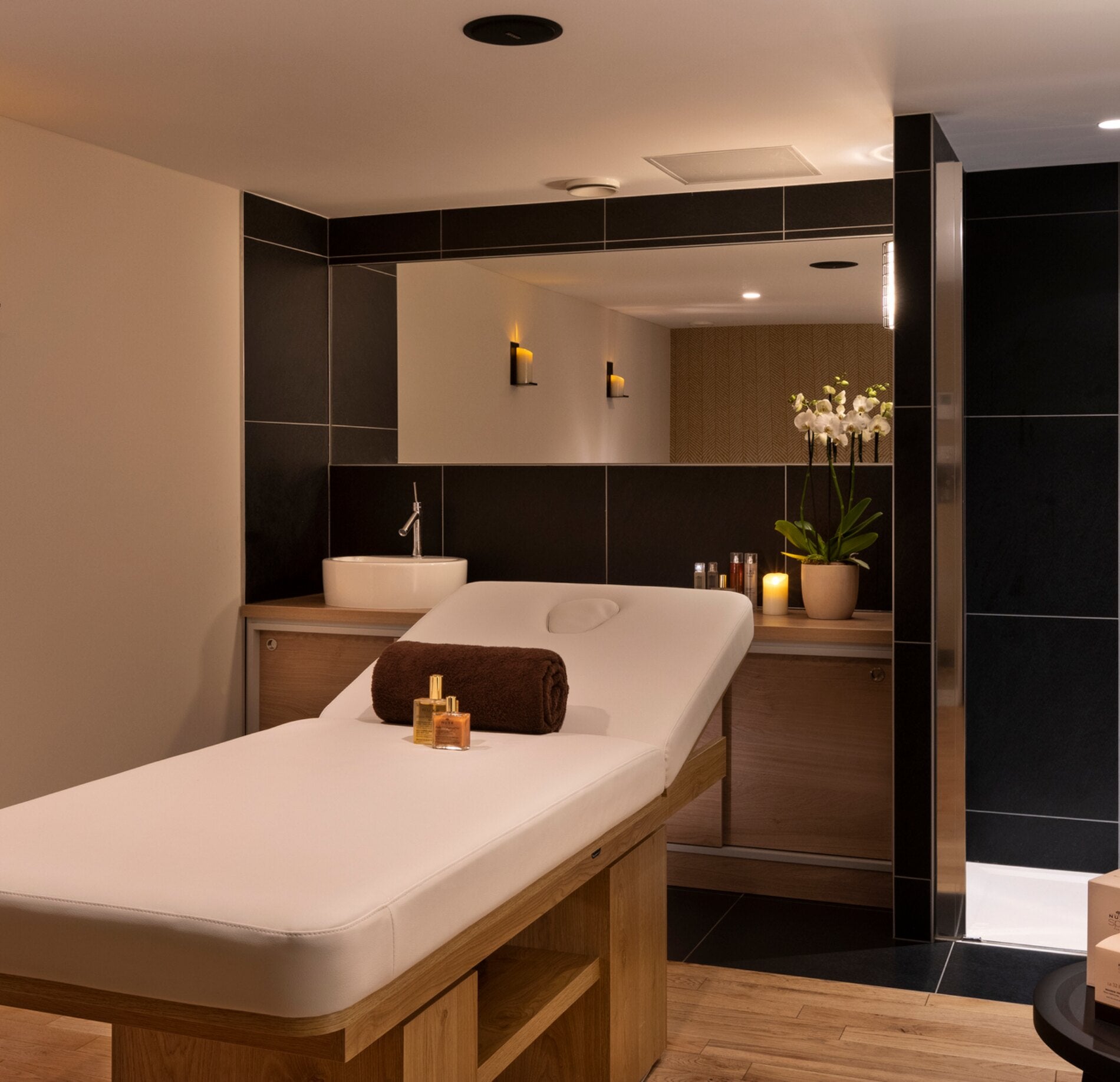 Une cabine de soin élégante du St'Alban Hotel & Spa, avec une table de massage recouverte d’un drap blanc, accompagnée de serviettes pliées et de flacons disposés avec soin.
Le décor associe des tons bois et noirs, un large miroir mural et un éclairage discret qui crée une atmosphère intime et relaxante.
