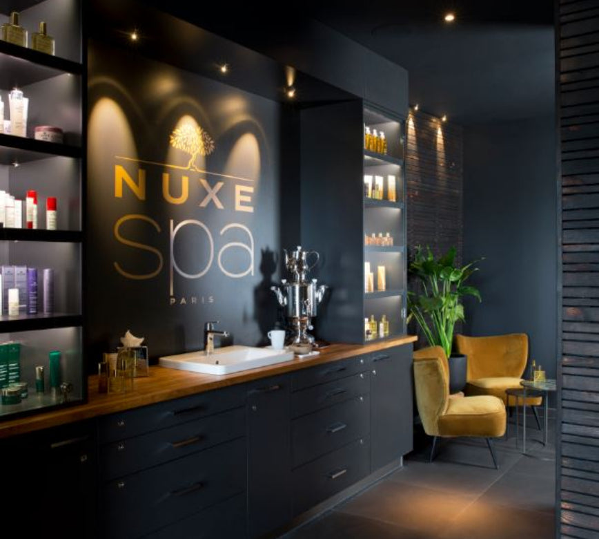 Un espace d’accueil élégant du Spa NUXE Terrass'Hotel, avec un comptoir en bois surmonté d’un lavabo et entouré d’étagères présentant des produits de soin.
Le mur noir met en valeur le logo lumineux “NUXE Spa Paris”, tandis que des fauteuils en velours et une plante verte apportent une touche chaleureuse et raffinée.