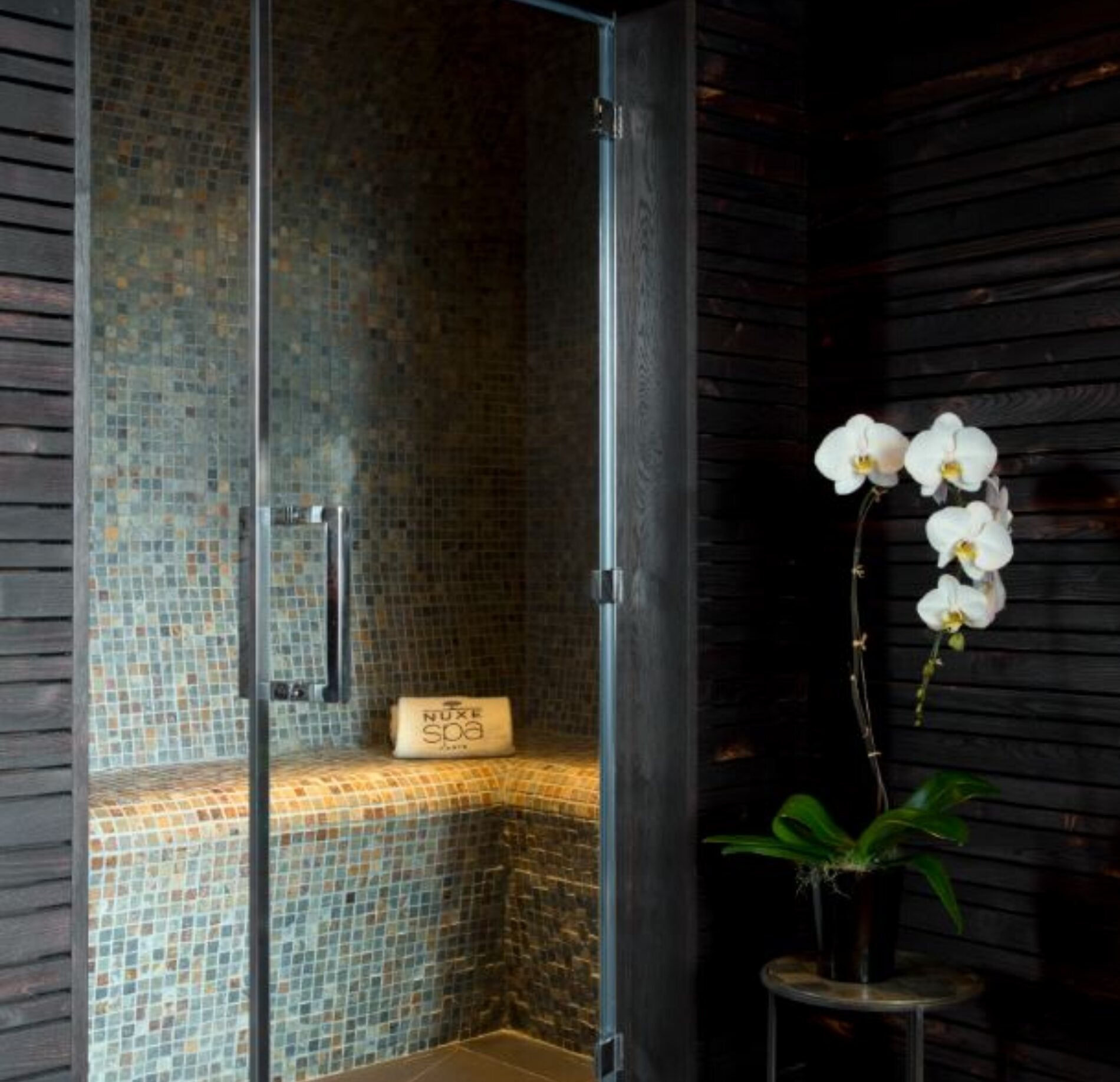 Un hammam élégant du Spa NUXE Terrass'Hotel, avec une cabine vitrée aux parois recouvertes de mosaïques aux tons chauds et une assise en angle.
À côté, une orchidée blanche posée sur une petite table et des murs en bois sombre créent une ambiance intime et sophistiquée propice à la détente.