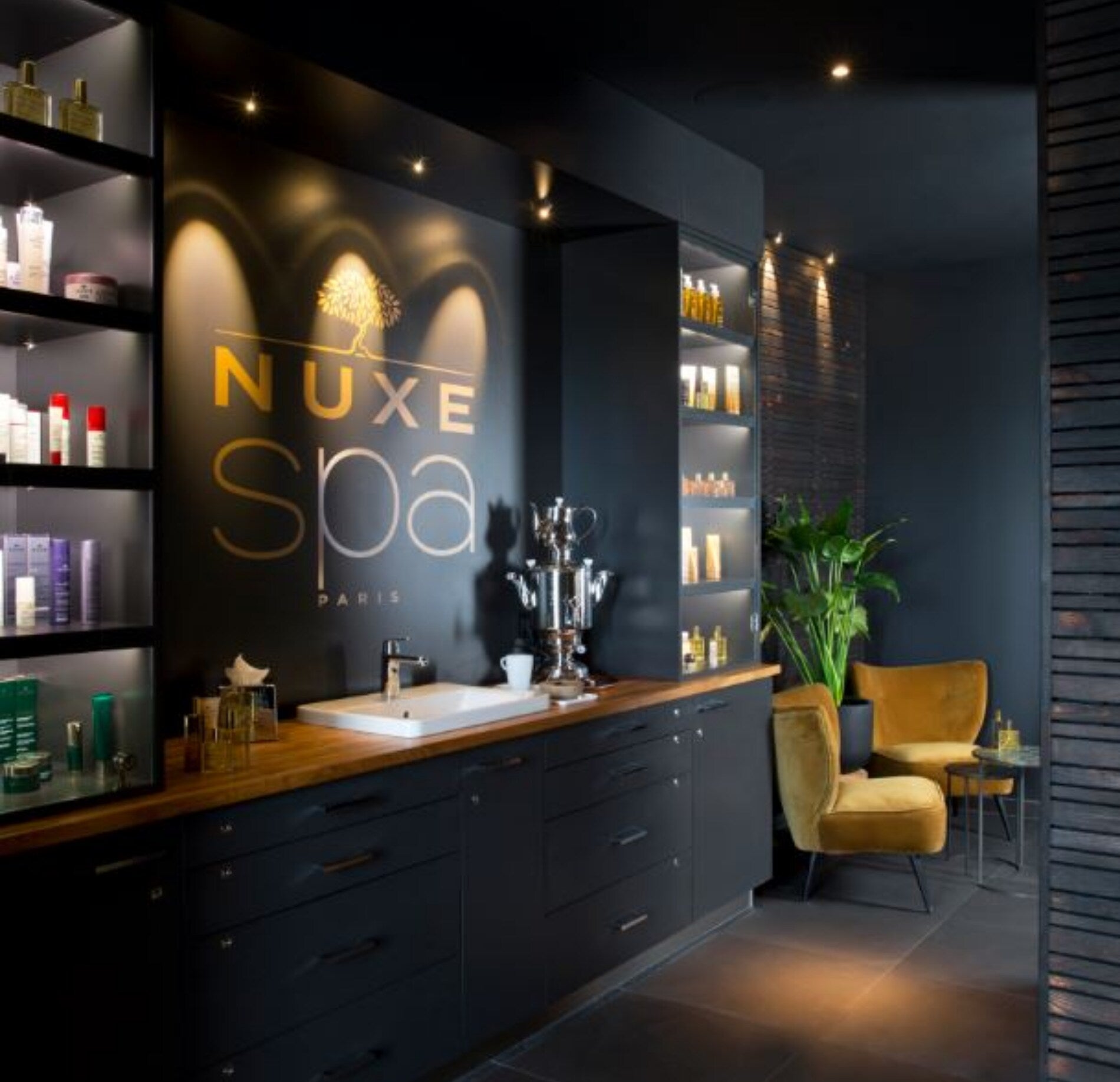 Un espace d’accueil élégant du Spa NUXE Terrass'Hotel, avec un comptoir en bois surmonté d’un lavabo et entouré d’étagères présentant des produits de beauté.
Le mur sombre met en valeur le logo lumineux “NUXE Spa Paris”, tandis que des fauteuils en velours et une plante verte apportent une ambiance chaleureuse et sophistiquée.
