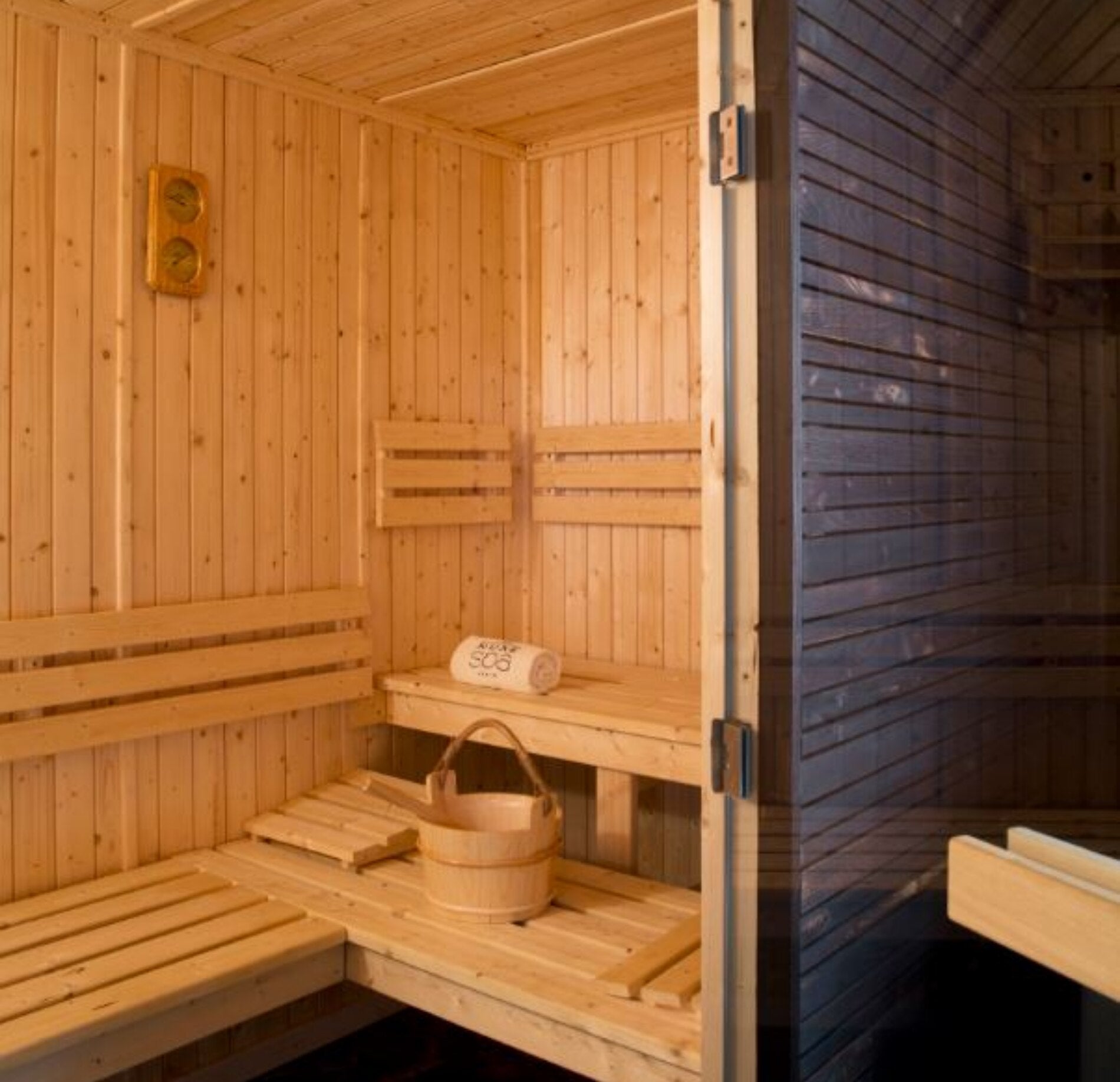 Un sauna en bois lumineux du Spa NUXE Terrass'Hotel, avec des bancs en lattes claires disposés sur plusieurs niveaux et un seau en bois accompagné d’une louche.
Les parois et le plafond en bois diffusent une chaleur visuelle apaisante, renforcée par un éclairage doux et des accessoires soigneusement disposés.