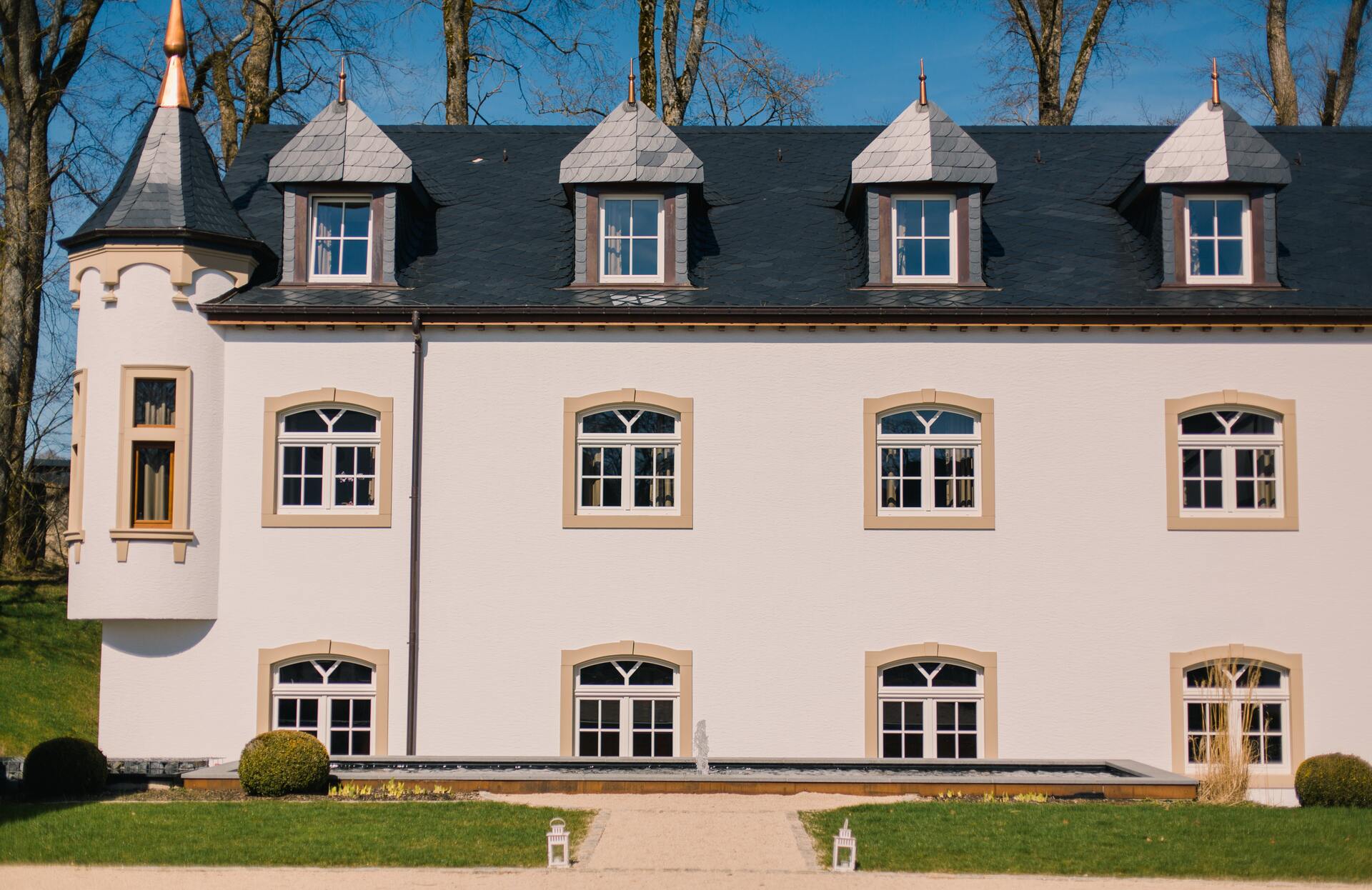 Une élégante bâtisse su spa Urspelt aux lignes classiques se dresse au cœur d’un jardin parfaitement entretenu, avec une façade claire rythmée par des fenêtres symétriques et des toits en ardoise.
Les détails architecturaux raffinés, la tourelle latérale et le chemin d’accès encadré de verdure offrent une première impression prestigieuse, évoquant un lieu d’exception dédié à la sérénité.