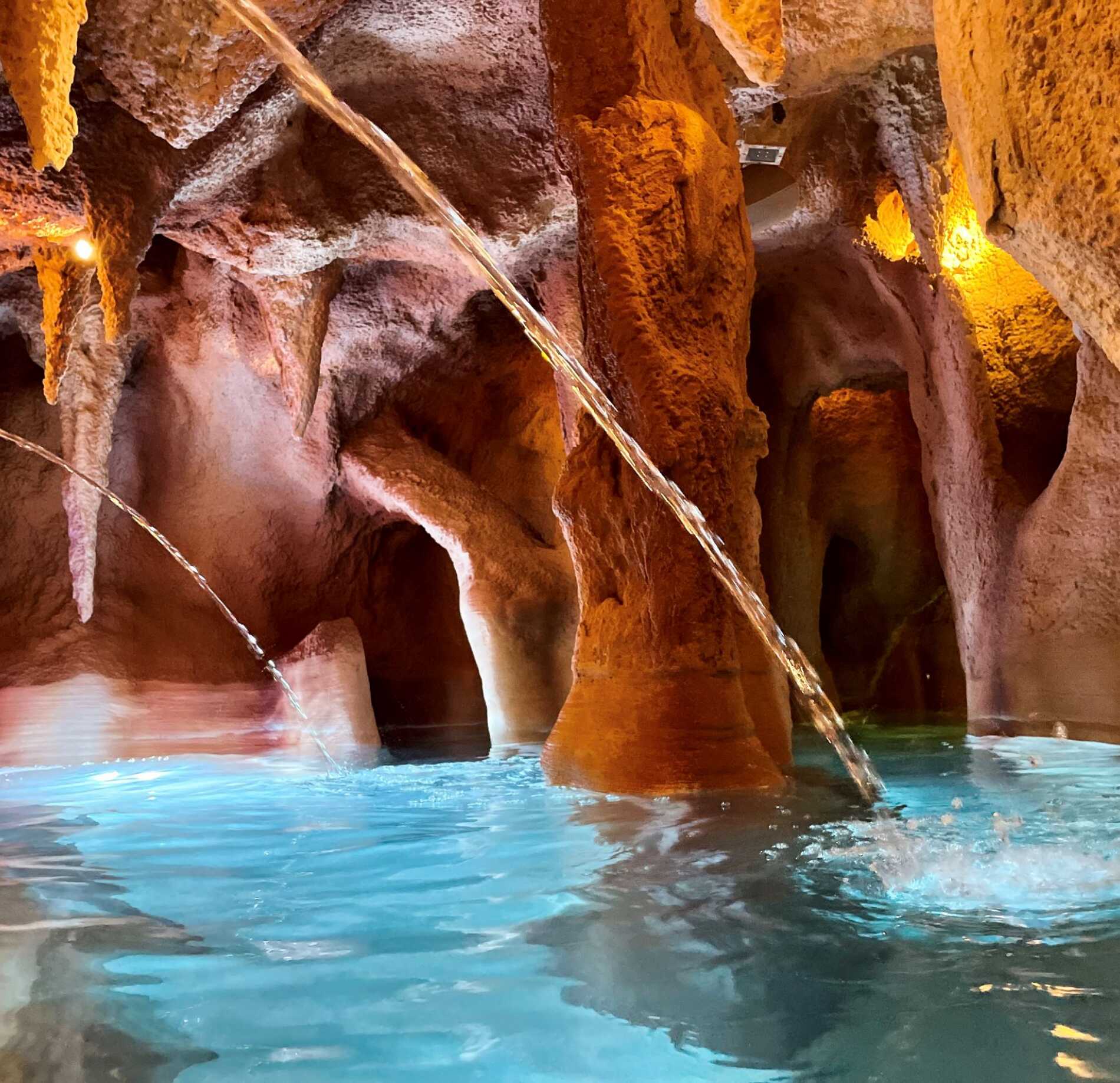 Espace aquatique spectaculaire du spa NUXE, inspiré d’une grotte naturelle, avec formations rocheuses sculptées, eau turquoise apaisante et jets d’eau en cascade, sublimé par un éclairage chaud créant une atmosphère enveloppante et dépaysante. 