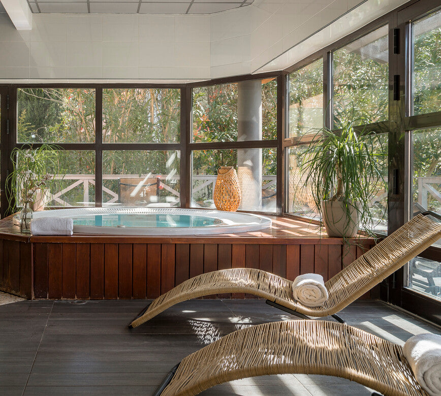 Coin bien-être du spa NUXE à l’Hôtel Valescure, composé d’un bain à remous encadré de bois, d’un transat ergonomique en matière tressée, de plantes décoratives et de larges fenêtres laissant entrer une abondante lumière naturelle.