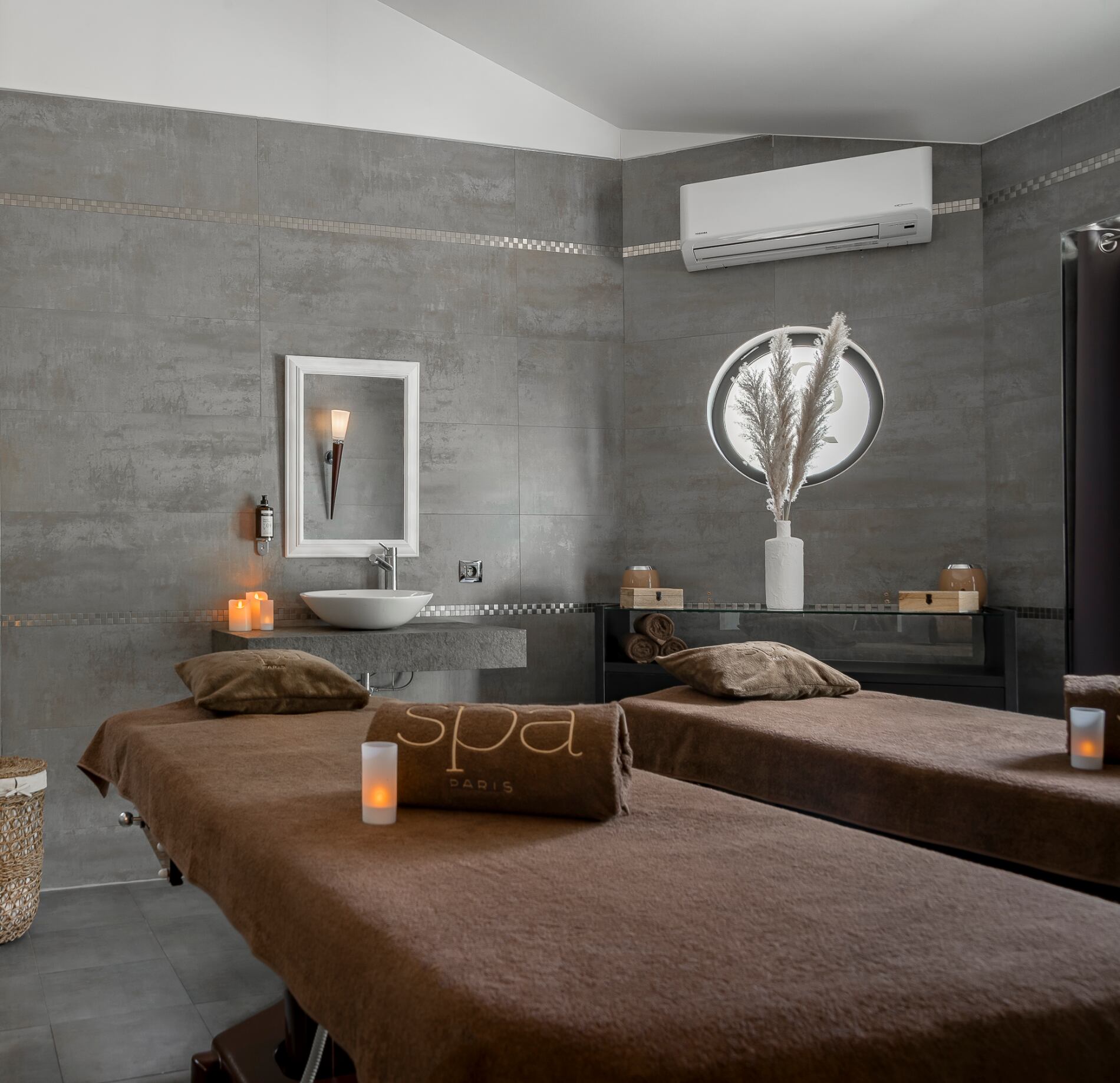 Cabine de soin élégante du spa NUXE à l’Hôtel Valescure, avec deux tables de massage habillées de linge brun, éclairage tamisé, bougies disposées avec soin et décoration minérale raffinée créant une atmosphère apaisante et haut de gamme.