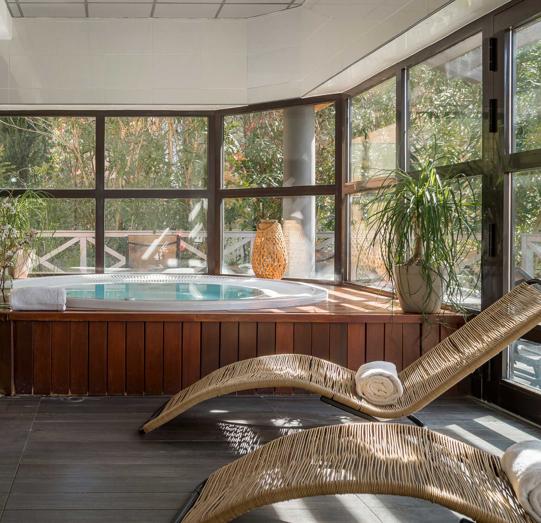 Espace détente lumineux du spa NUXE à l’Hôtel Valescure, avec jacuzzi encastré entouré de bois chaleureux, transat en fibres naturelles et grandes baies vitrées donnant sur une végétation verdoyante, créant une atmosphère apaisante et raffinée.