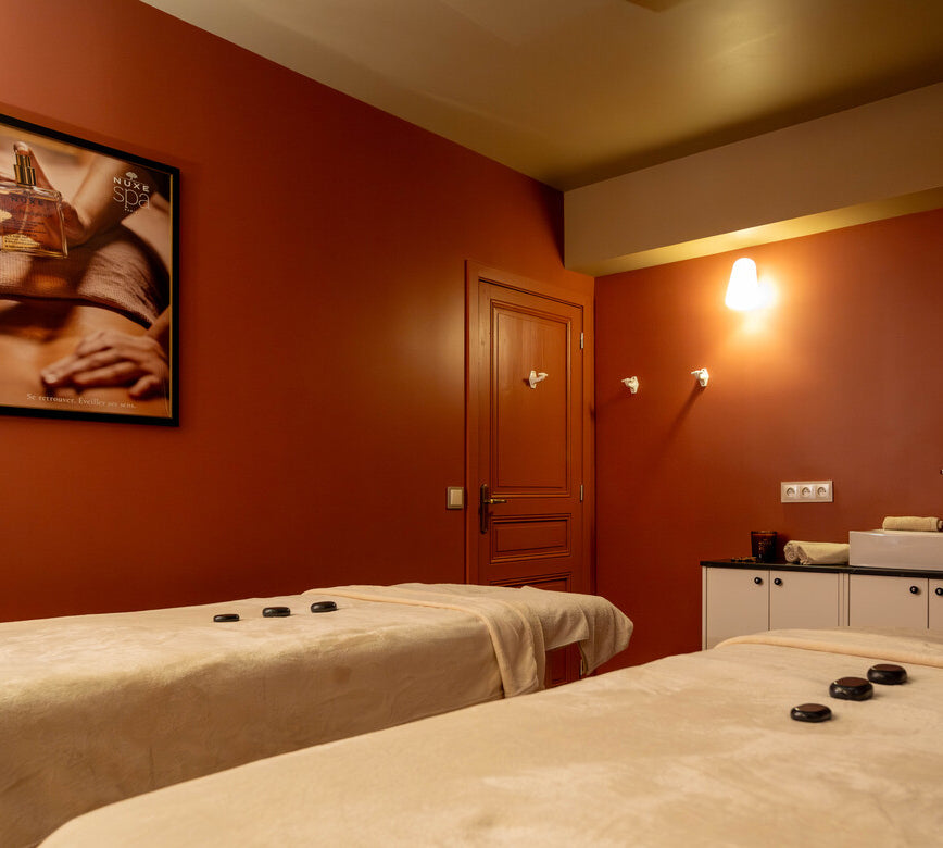 Salle de soins cosy et raffinée au spa NUXE, habillée de tons chauds, avec tables de massage soigneusement préparées, lumière tamisée et décoration épurée.