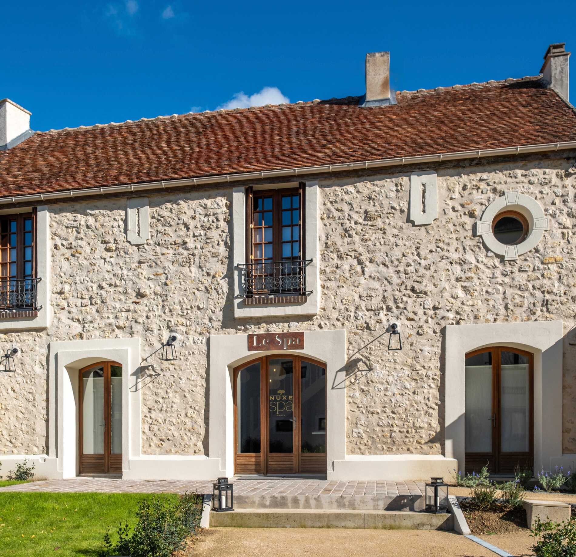 Façade authentique en pierre du spa NUXE Les Varennes, sublimée par une architecture traditionnelle aux tons clairs, avec portes en bois élégantes et toit en tuiles, sous un ciel bleu lumineux créant une atmosphère accueillante et raffinée.