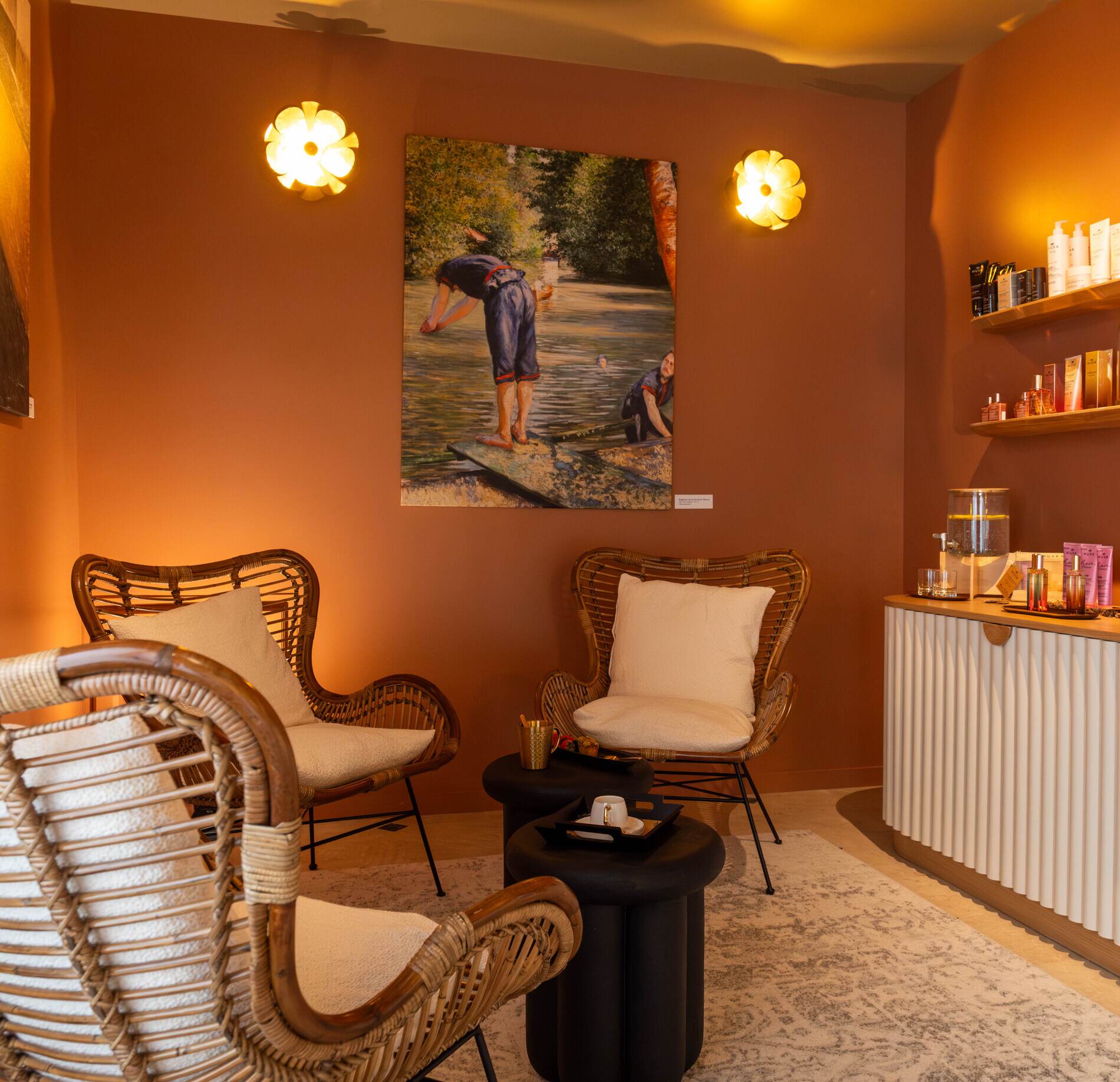 Espace d’accueil cosy et raffiné du spa NUXE, aux murs aux teintes chaleureuses, avec fauteuils en fibres naturelles, éclairage doux et étagères de produits, créant une atmosphère élégante et apaisante propice à la détente. 
