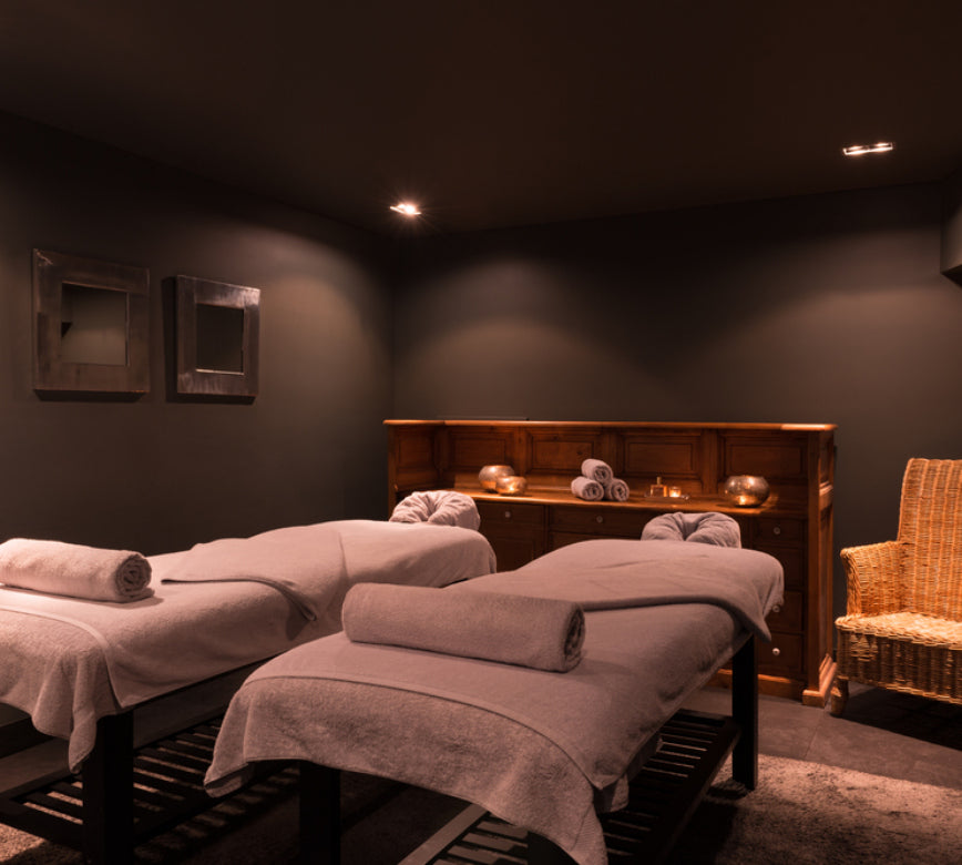 Une cabine de soin chaleureuse de l'Hotel Spa Vent d'Ouest, avec deux tables de massage recouvertes de draps épais et surmontées de serviettes soigneusement roulées.
Le décor intimiste, composé de murs sombres, de mobilier en bois et d’un éclairage tamisé, crée une atmosphère enveloppante propice à la détente.