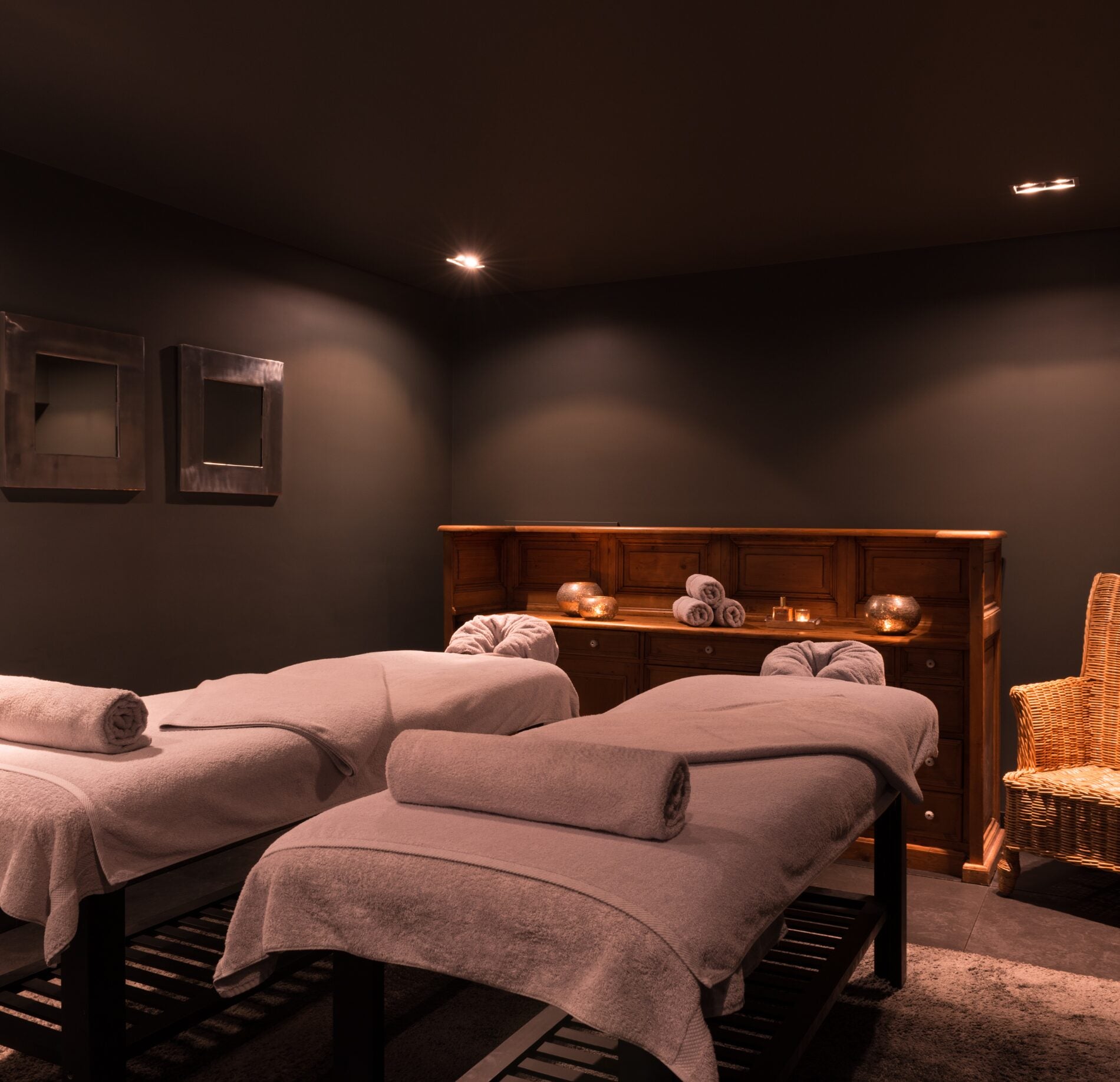 Une cabine de soin enveloppante de l'Hotel Spa Vent d'Ouest, avec deux tables de massage recouvertes de draps épais et de serviettes roulées soigneusement disposées.
Le mobilier en bois, les murs foncés et l’éclairage tamisé diffusé au plafond créent une atmosphère intime et chaleureuse dédiée à la détente.