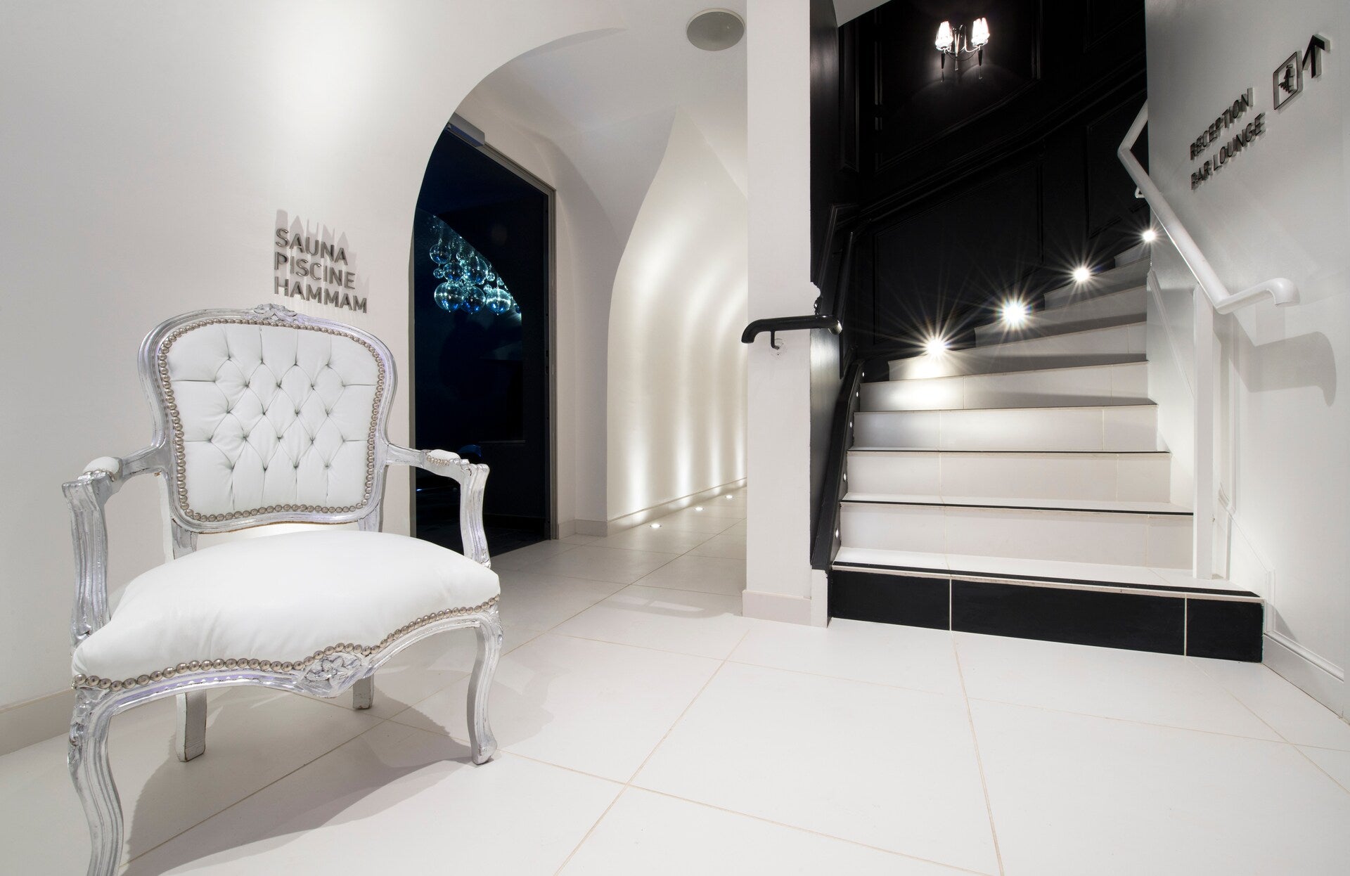 Une entrée élégante et épurée s’ouvre sur un espace bien‑être, avec un fauteuil capitonné blanc aux finitions travaillées posé au premier plan sur un sol clair immaculé.
À droite, un escalier noir et blanc subtilement éclairé mène à d’autres espaces, tandis qu’une arche douce révèle l’accès au sauna, à la piscine et au hammam dans une ambiance sophistiquée et haut de gamme.