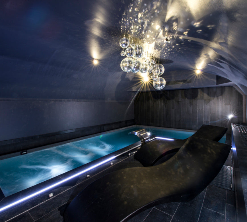 Un espace bien‑être raffiné de l'Hotel Spa Vertigo, avec une piscine intérieure aux courbes modernes et une eau turquoise animée de remous apaisants.
Au premier plan, des transats ergonomiques sombres font face au bassin, tandis qu’un lustre suspendu composé de sphères lumineuses diffuse des reflets élégants au plafond.