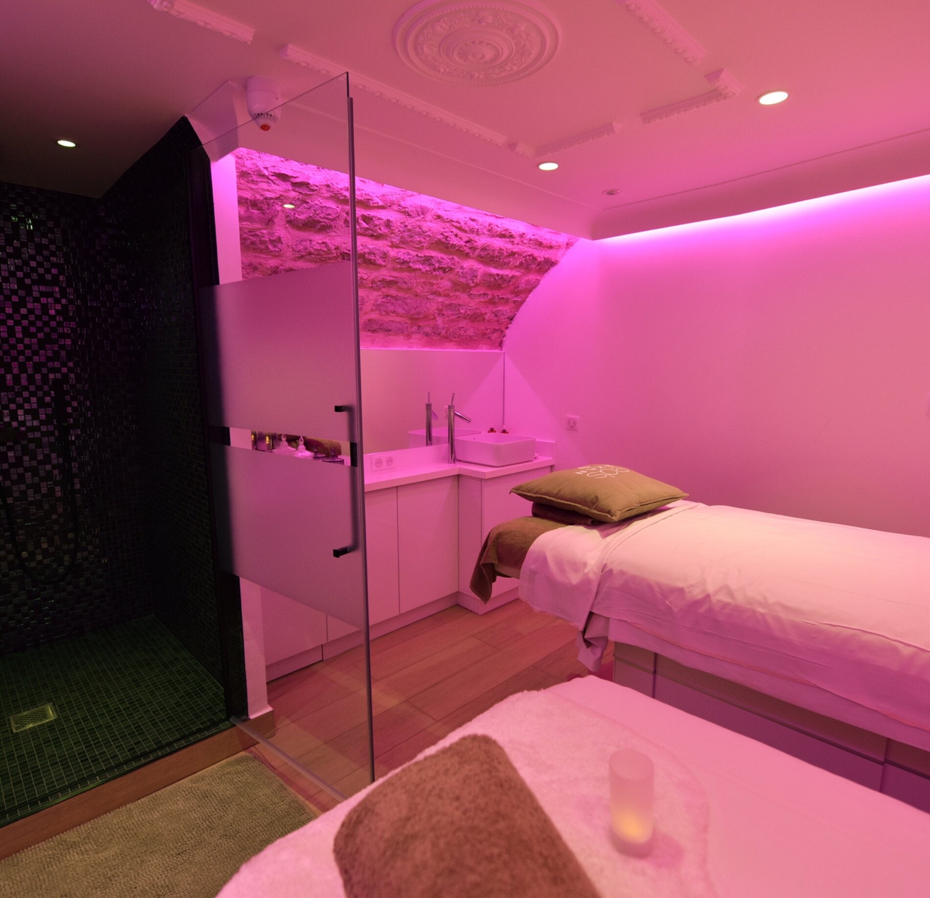 Une cabine de soin contemporaine de l'Hotel Spa Vertigo, avec une table de massage recouverte de linge clair et un coussin posé avec soin au centre.
À l’arrière, une douche vitrée et un mur en pierre éclairé par une lumière rose diffuse créent une ambiance douce et immersive, mêlant modernité et chaleur.