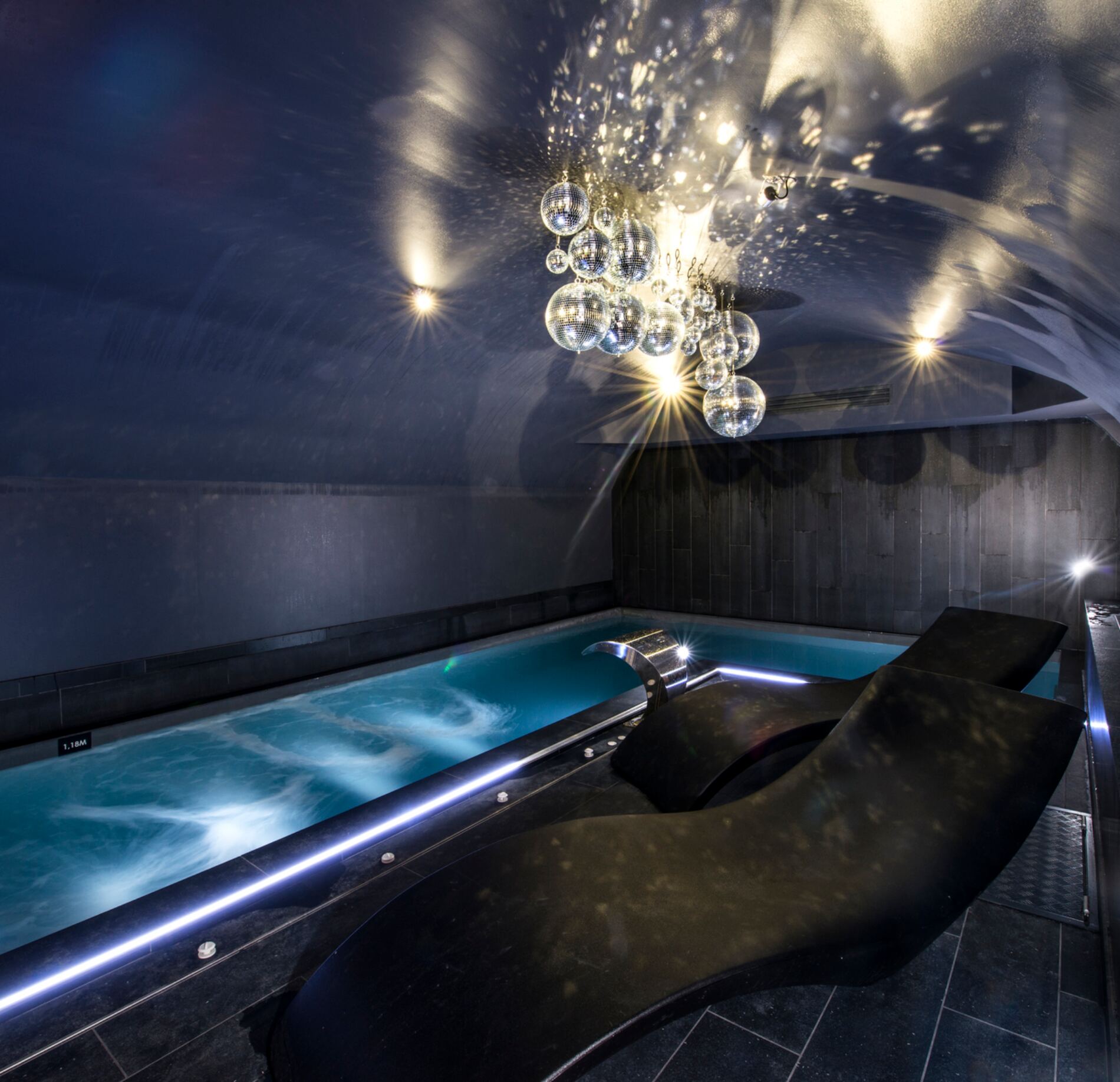 Un espace bien‑être raffiné de l'Hotel Spa Vertigo, avec une piscine intérieure aux lignes modernes et une eau turquoise animée de remous relaxants.
Au premier plan, des transats ergonomiques sombres font face au bassin, tandis qu’un lustre suspendu composé de bulles lumineuses diffuse des reflets sophistiqués au plafond.
