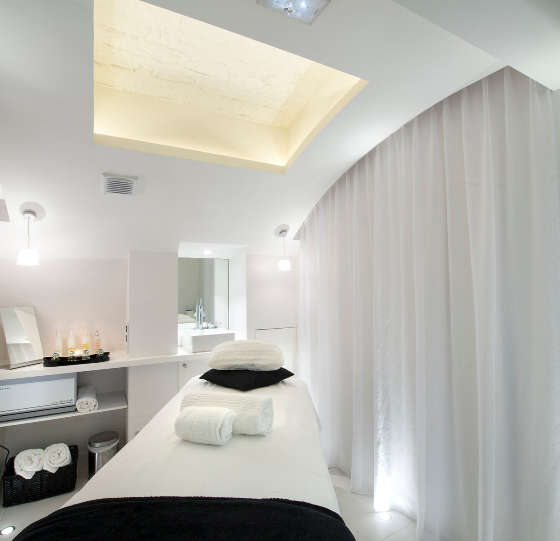 Une cabine de soin lumineuse de l'Hotel Spa Vertigo, avec une table de massage recouverte de linge blanc et des serviettes pliées avec précision au centre.
L’espace épuré, aux tons clairs et aux rideaux fluides, est sublimé par un éclairage doux venant du plafond, créant une atmosphère apaisante et raffinée.